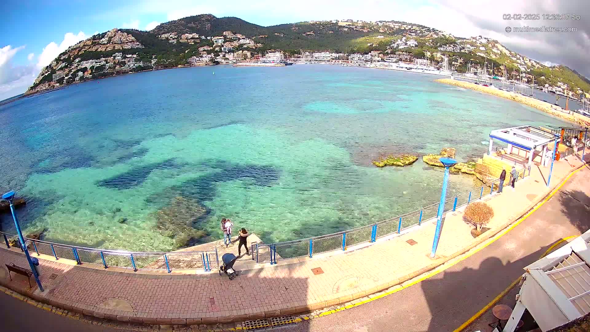 A clear, sunny day reveals a calm, shallow turquoise sea lapping a paved waterfront walkway, with a hillside town and harbor in the background.