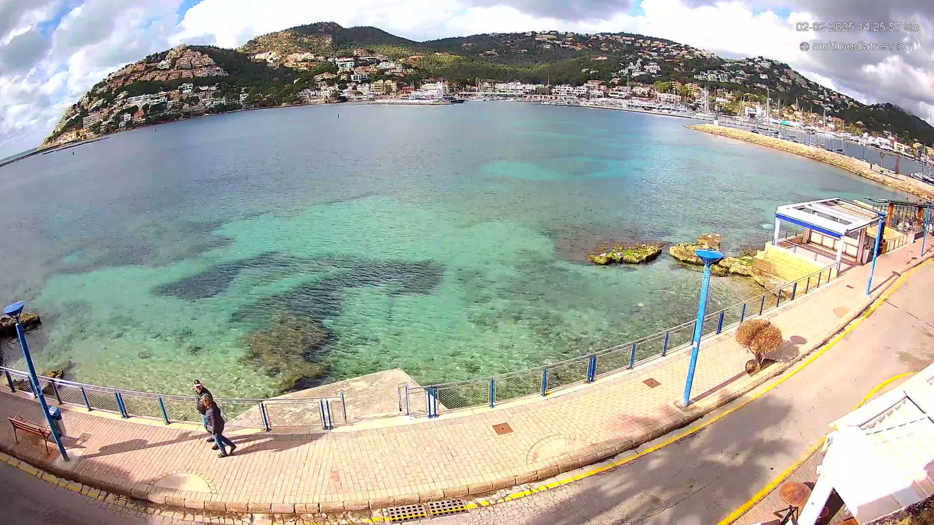 A partly cloudy day shows a clear, shallow, teal-colored bay with a paved walkway, a small building, and a hill covered in buildings in the background.