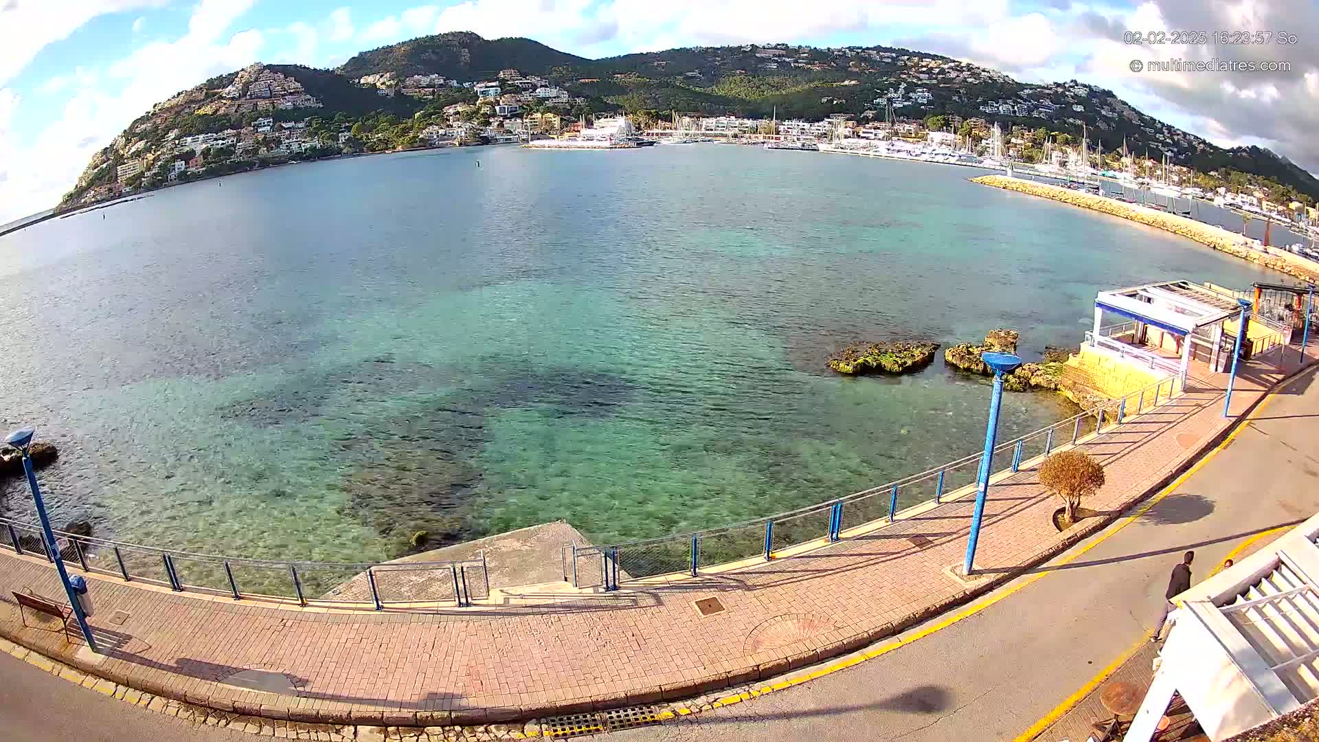 A paved walkway curves along a calm, clear, turquoise sea, with a hill covered in buildings and greenery in the background under a partly cloudy sky.