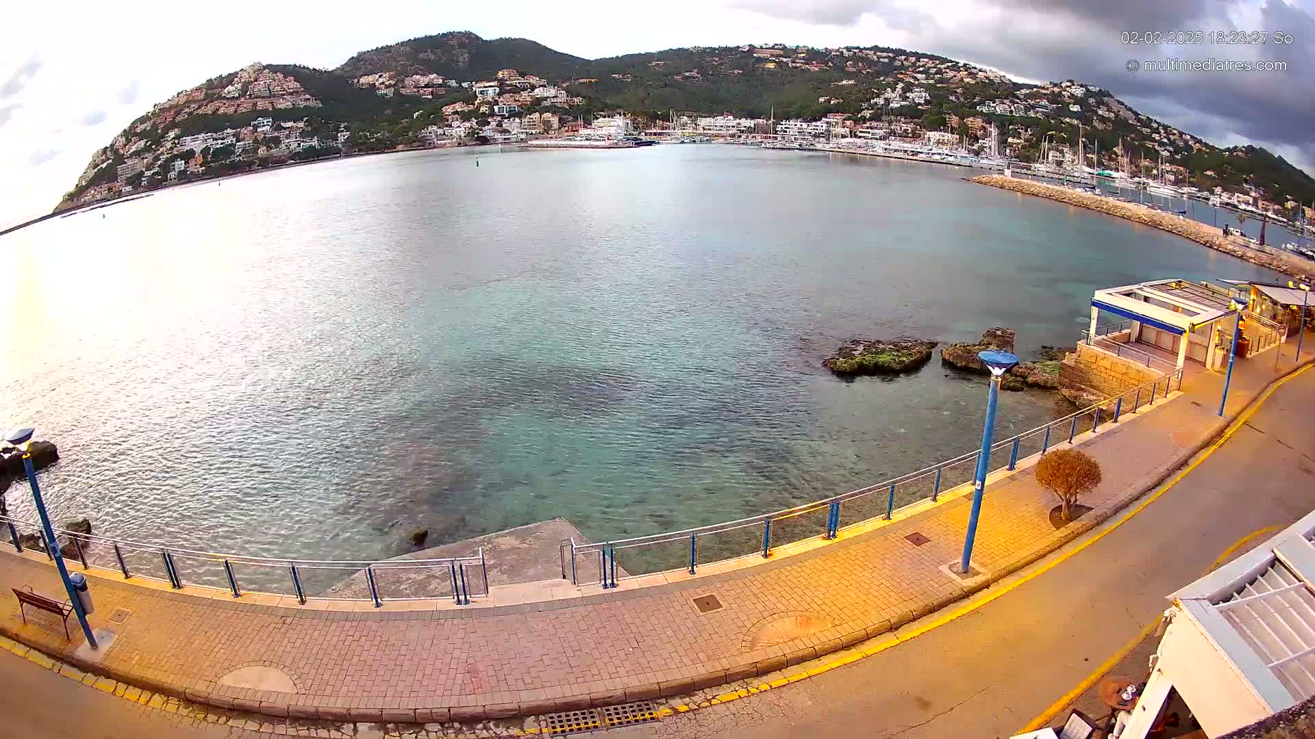 A seaside promenade overlooks a calm bay filled with boats, with a hill covered in buildings in the background under a cloudy sky.