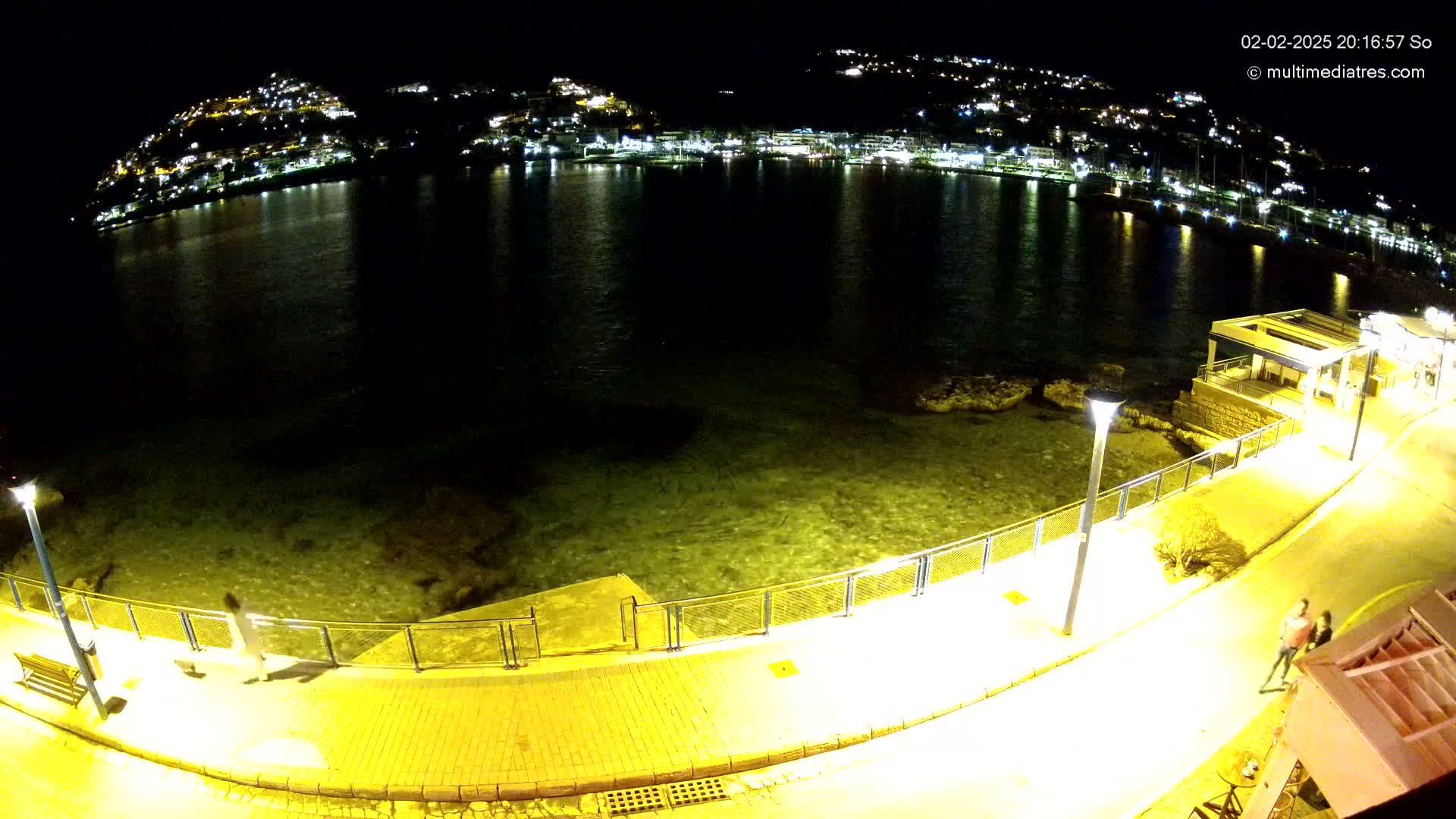 A nighttime view of a calm, lit waterfront with a paved walkway, a few people walking, and a hilltop town illuminated in the background.