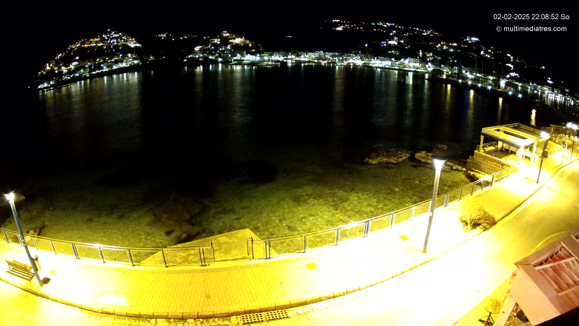 A nighttime view of a waterfront walkway illuminated by streetlights, overlooking a calm bay reflecting the lights of a town nestled on hillsides in the distance.