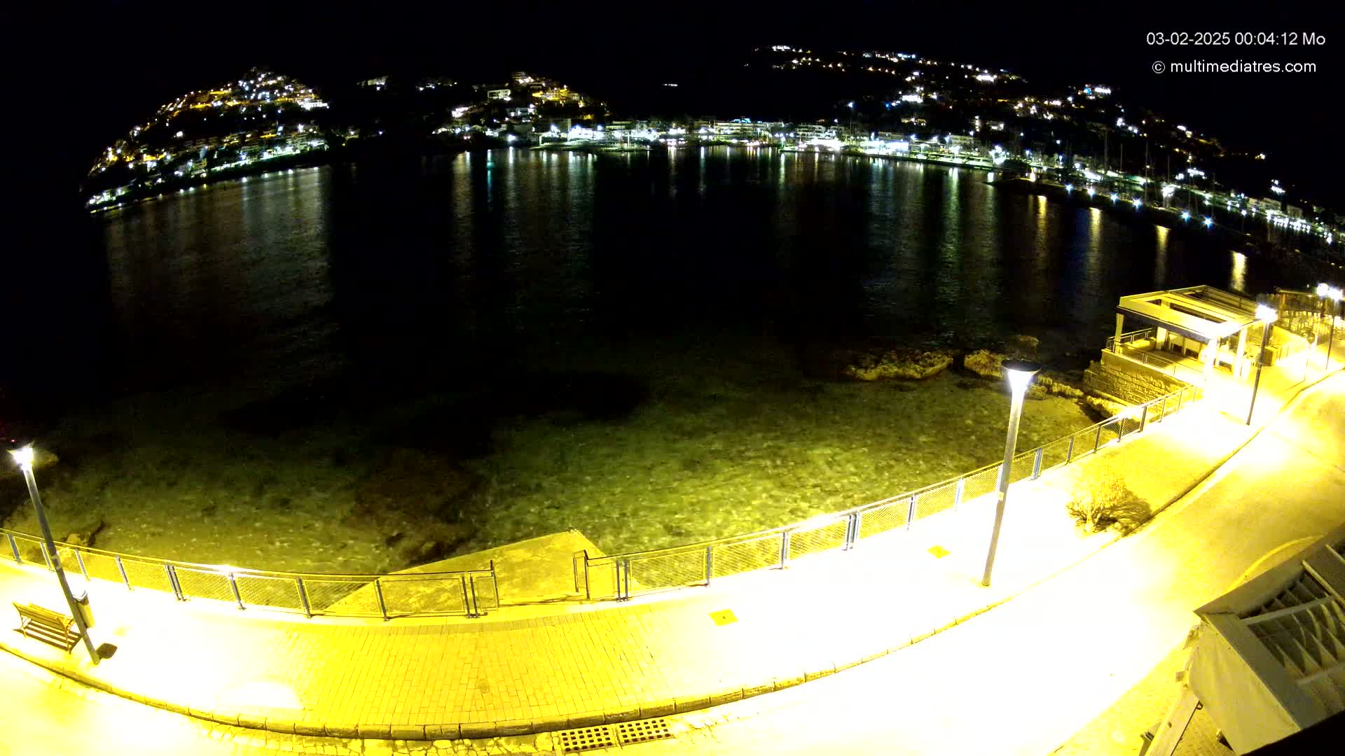 A nighttime view of a calm bay with a town's lights reflected in the water, seen from an elevated position on a paved walkway with lights along the shoreline.