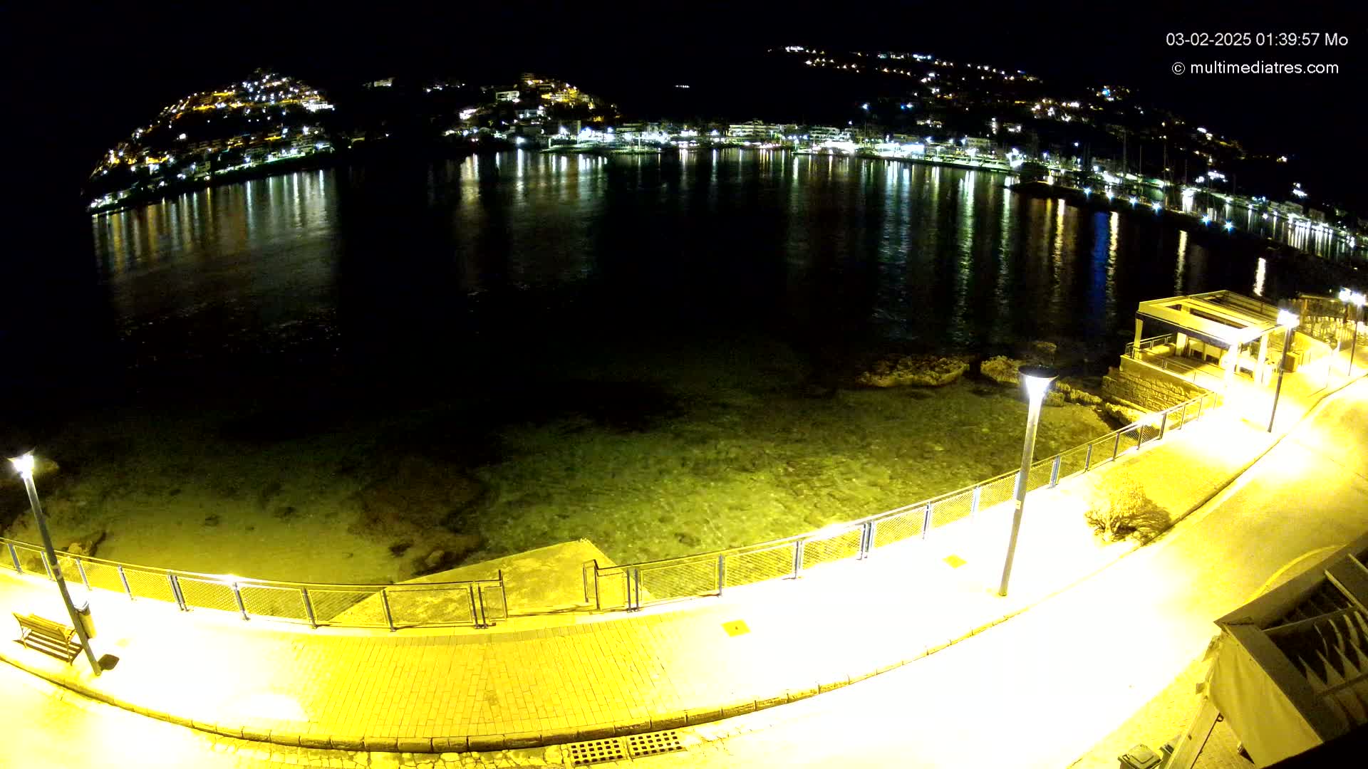 A nighttime, high-angle view shows a waterfront walkway illuminated by lamps, overlooking a calm body of water reflecting the lights of a town built on a hillside.