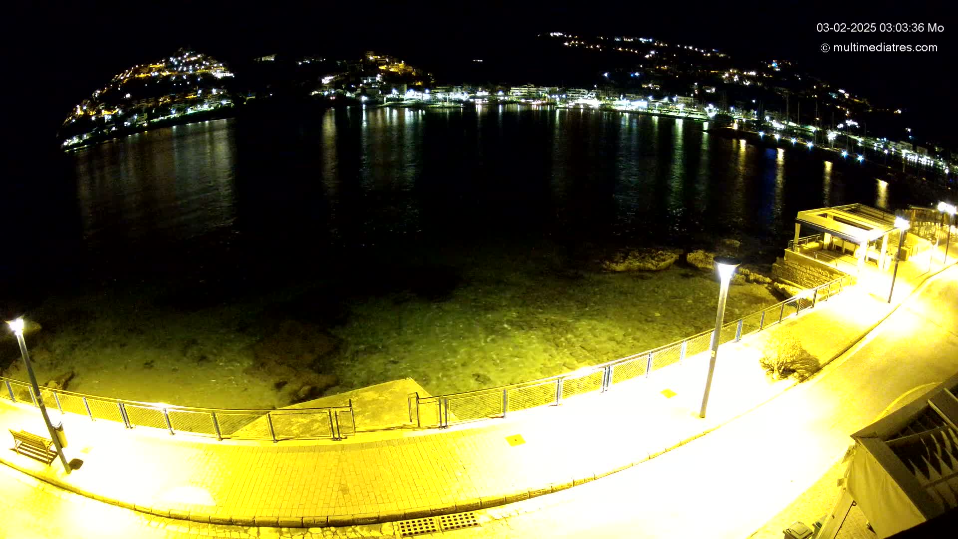 A nighttime, high-angle view shows a calm body of water reflecting the lights of a town built on a hill, with a paved waterfront path illuminated by lamps.