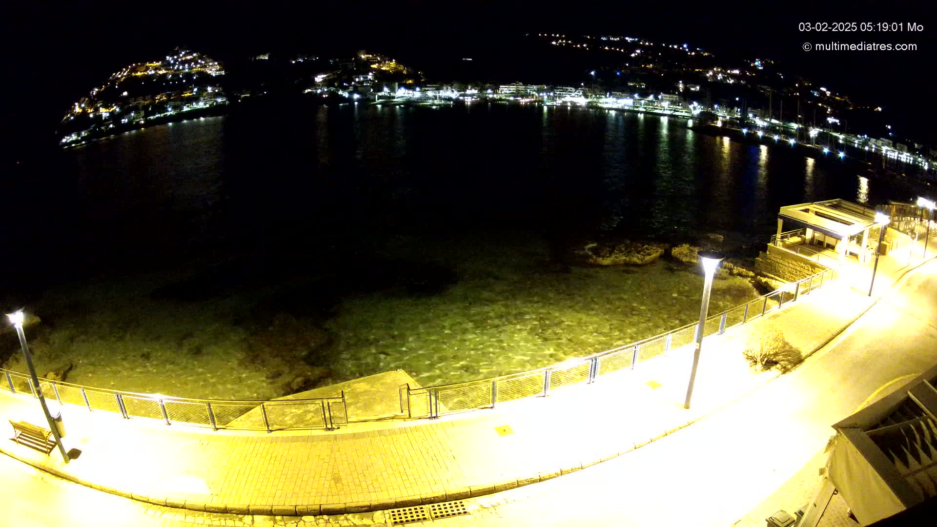 A nighttime view of a calm, shallow body of water next to a paved walkway lined with lights, overlooking a town illuminated on a hill.