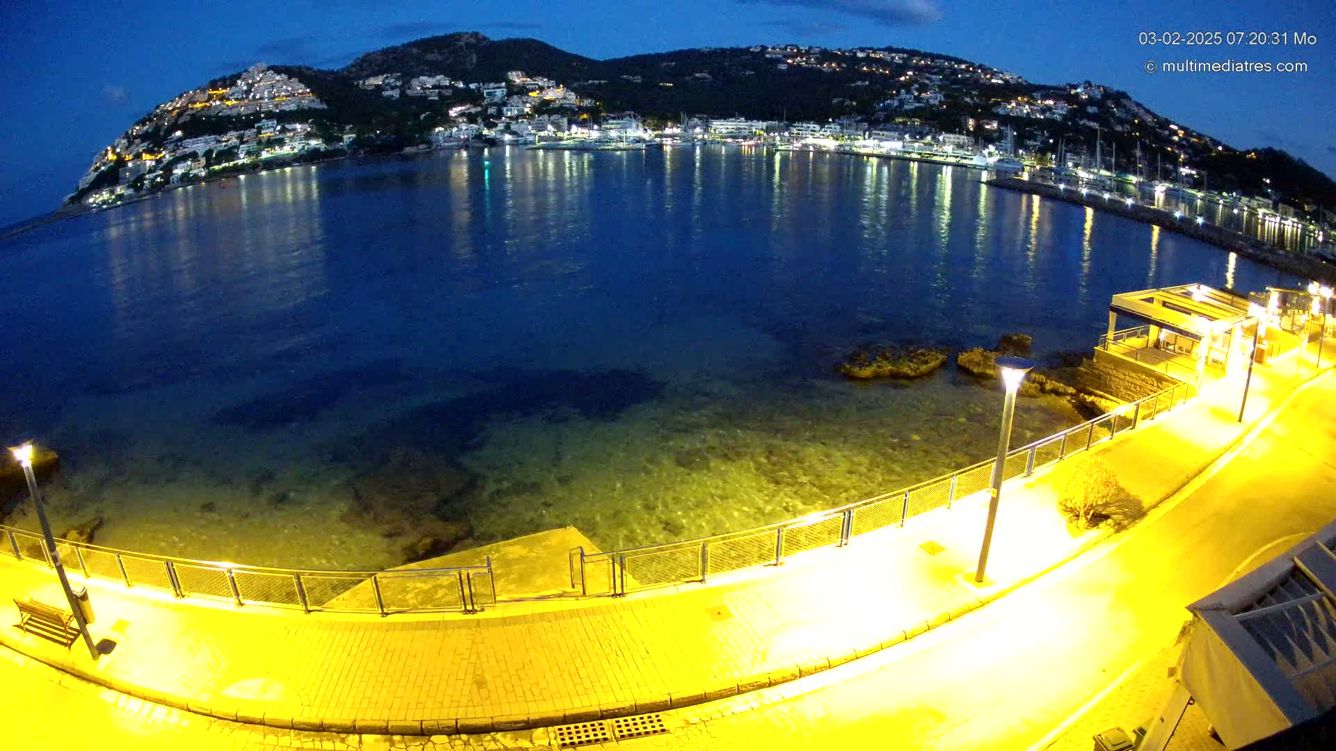 A nighttime coastal scene shows a paved walkway illuminated by streetlights next to a calm, dark-blue bay reflecting the lights of a town nestled on a hill in the distance.