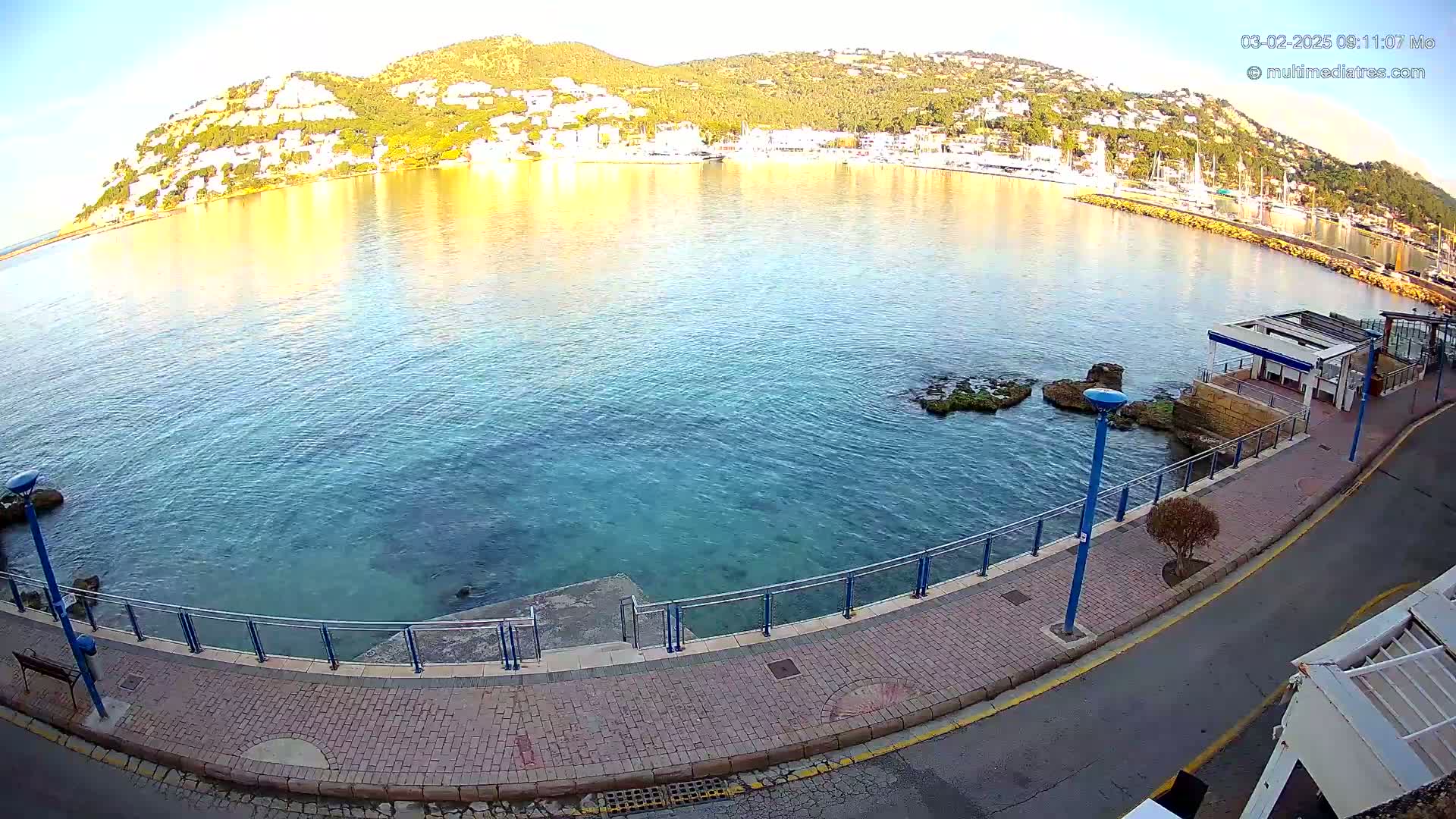 A calm, clear body of water laps against a paved waterfront walkway, with a hill dotted with white buildings in the background under a bright, sunny sky.