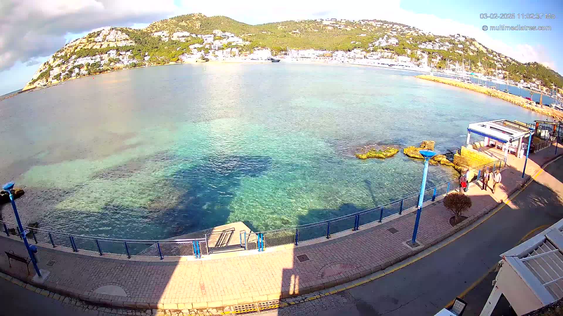 A seaside boardwalk with clear, shallow turquoise water and a hill covered in white buildings in the background under a partly cloudy sky.