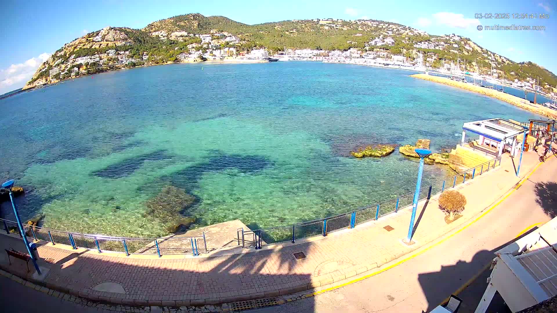 Clear, shallow, turquoise water laps a paved waterfront path next to a small, hilly town under a sunny sky.