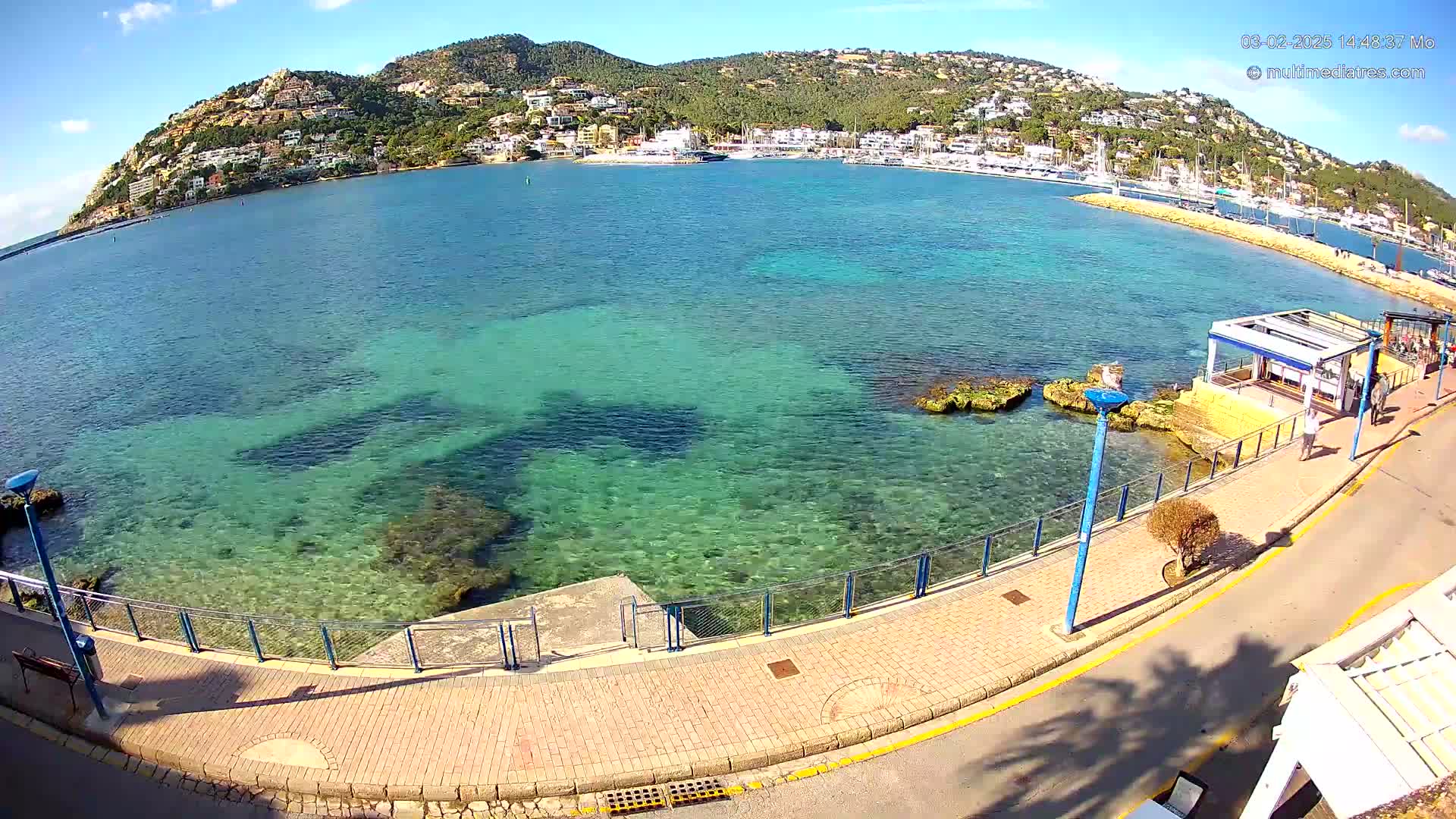 A paved walkway borders a calm, clear turquoise bay with a hilltop town in the background under a sunny sky.