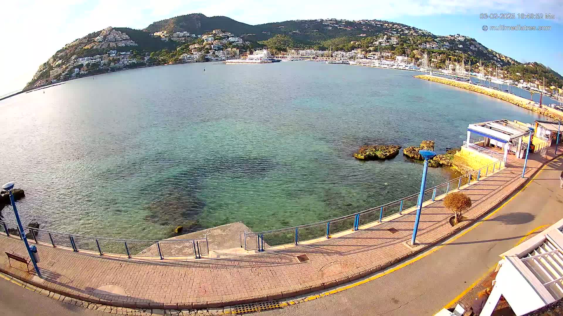 A calm, clear body of water with a marina filled with boats is seen from a high vantage point, next to a paved walkway, with a hill covered in buildings in the background under a sunny sky.