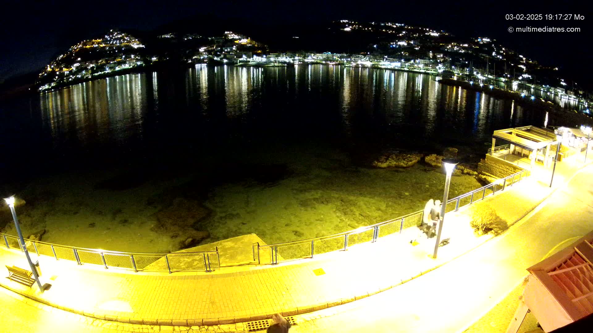 A nighttime view of a calm body of water reflecting the lights of a hillside town, with a paved walkway and railings along the shore.