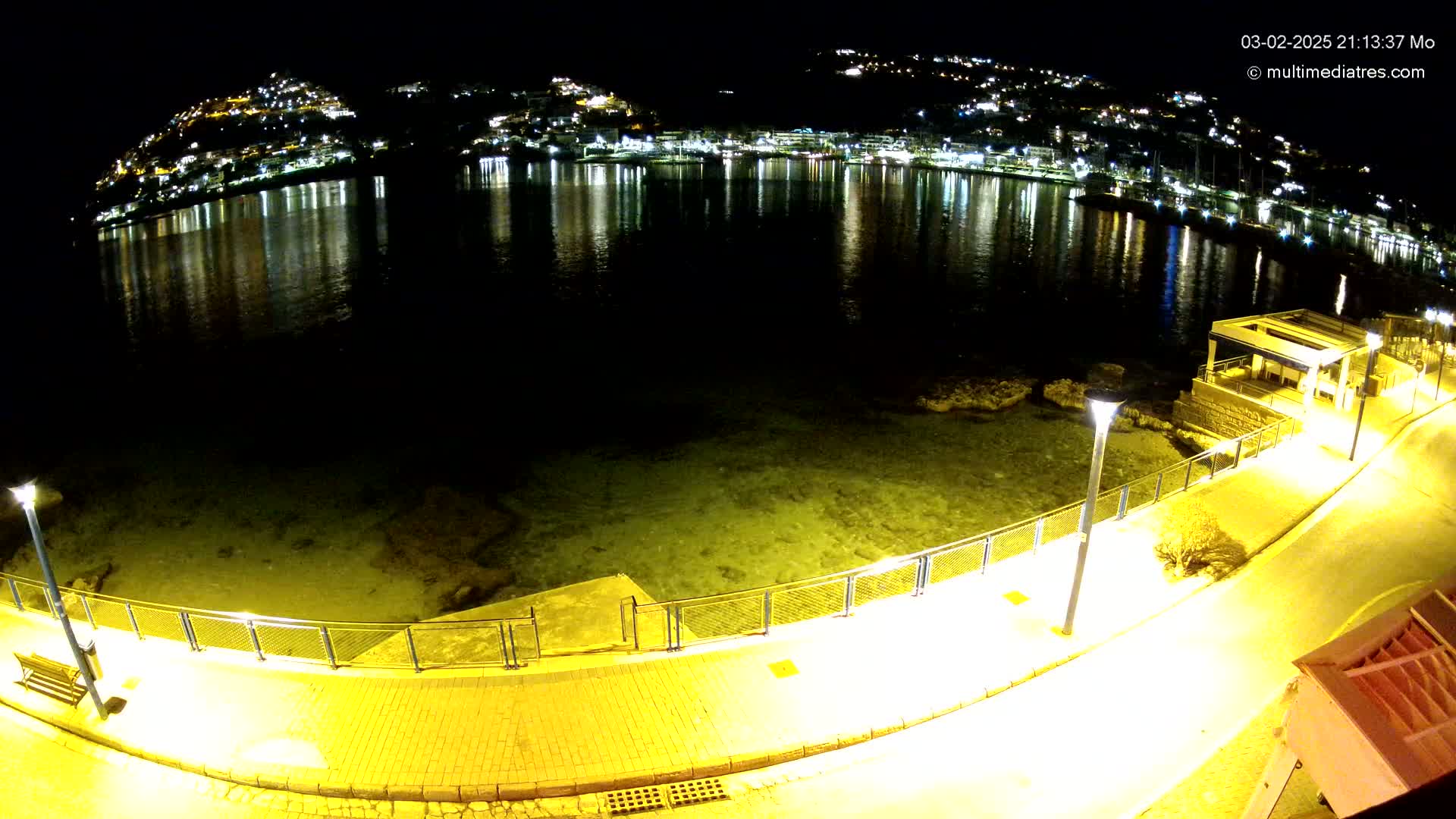 A nighttime view of a calm bay reflecting the lights of a town on its shores, seen from a paved walkway with lights illuminating the scene.