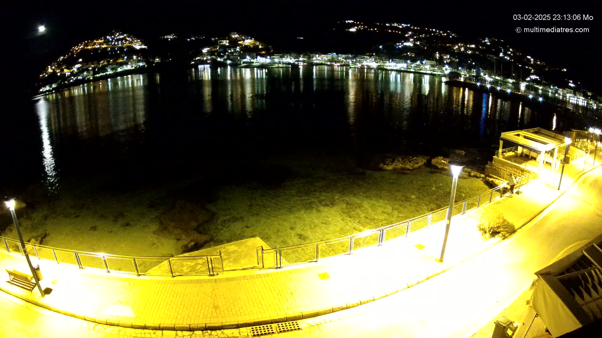 A moonlit night scene shows a paved walkway curving along a calm, shallow body of water, with a hill of illuminated buildings reflected in the water.