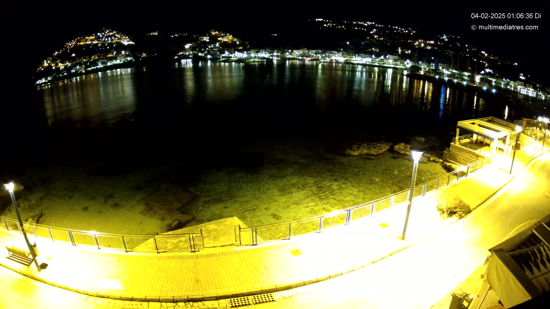 A nighttime, overhead view shows a calm body of water reflecting the lights of a town nestled on hillsides, with a paved walkway and metal railing alongside the water's edge, illuminated by streetlights.