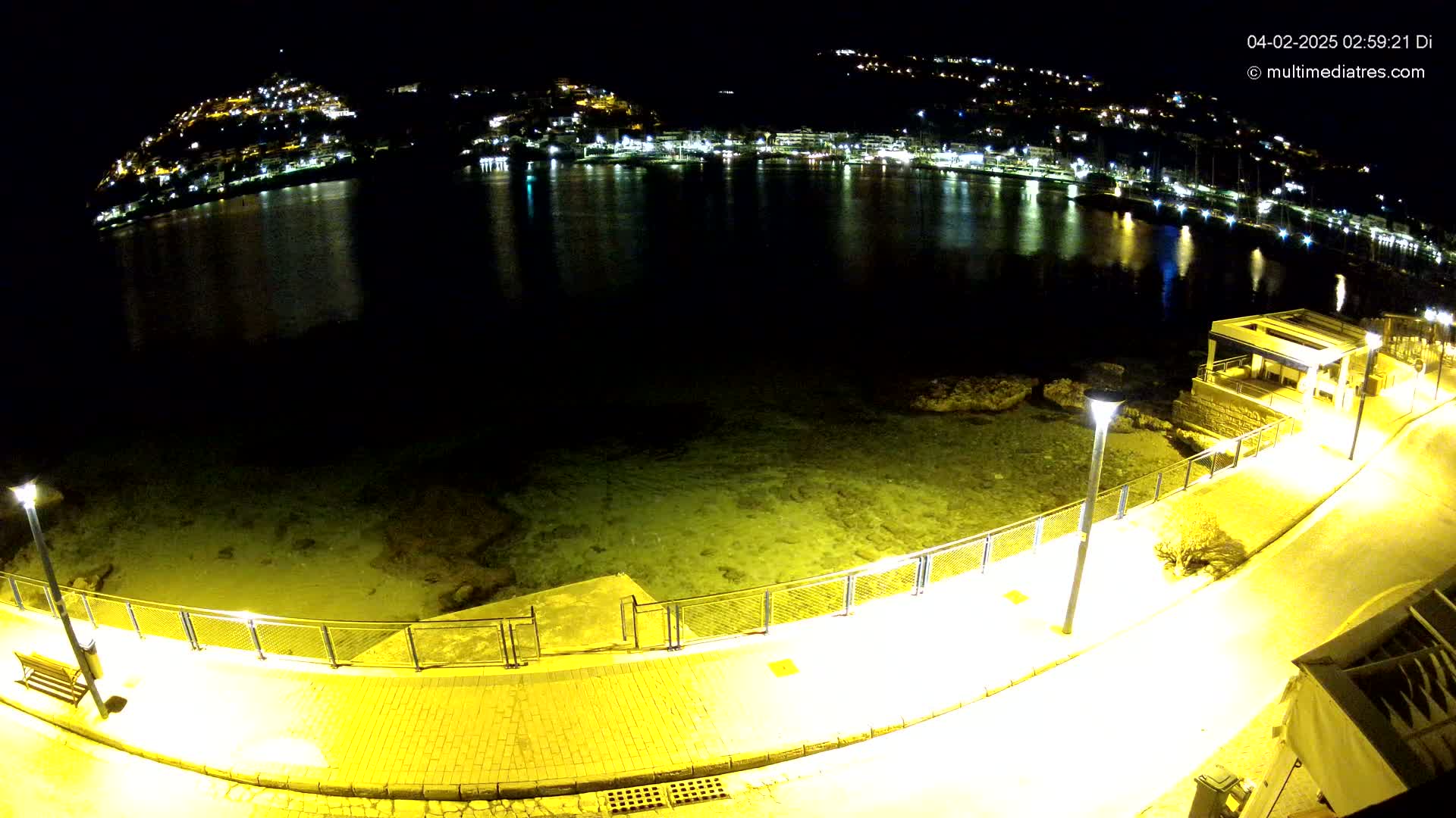 A nighttime view of a calm body of water reflecting the lights of a town on a hillside, with a paved walkway and lampposts lining the shore.