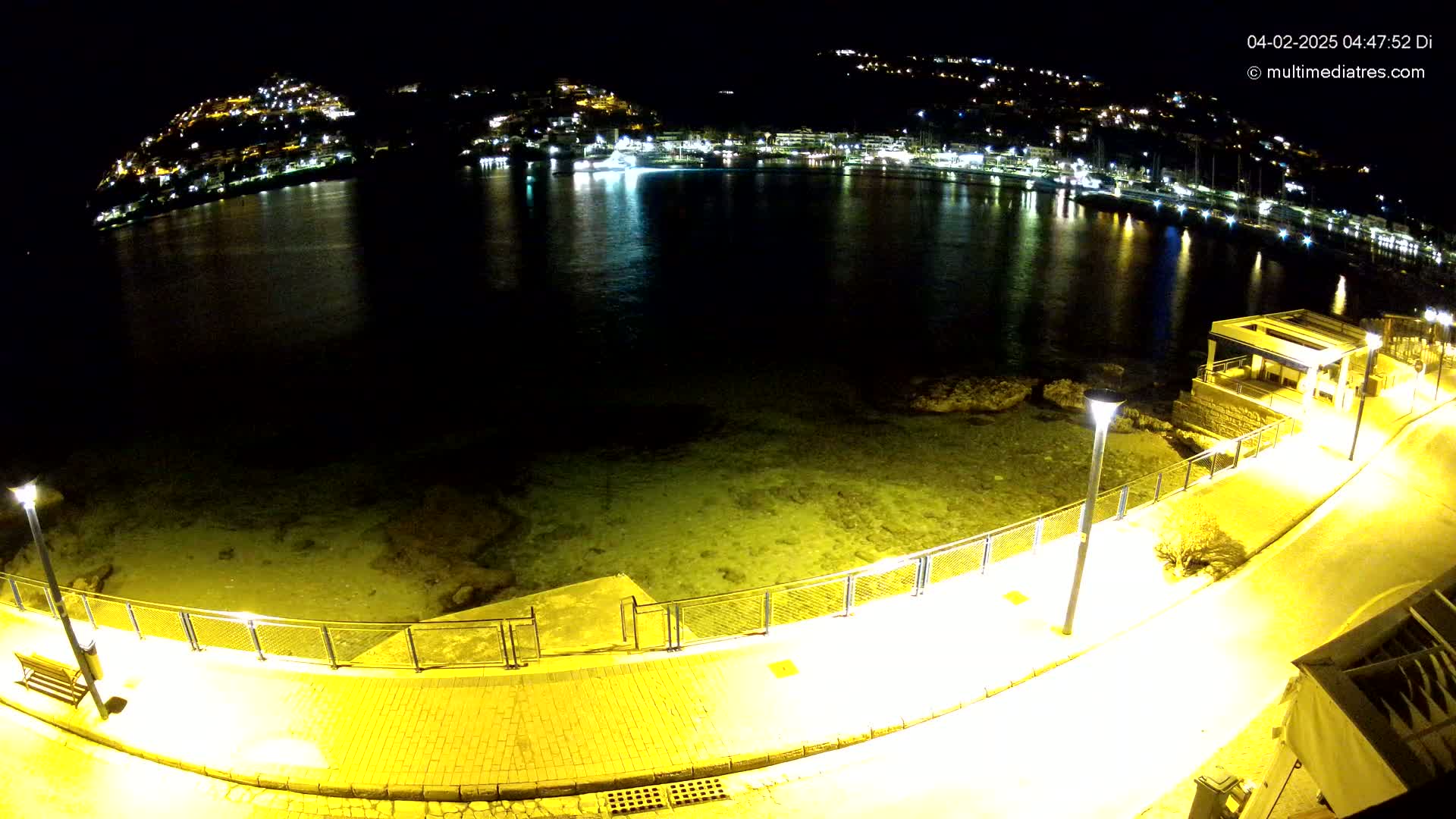 A nighttime, illuminated waterfront path curves along a calm sea, overlooking a town nestled on a hillside.