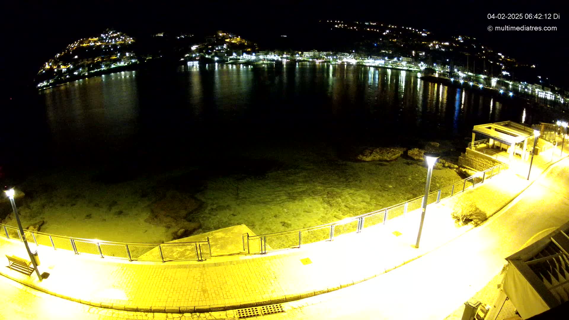 A nighttime, high-angle view shows a paved walkway alongside a calm body of water reflecting the lights of a town built on a hillside.