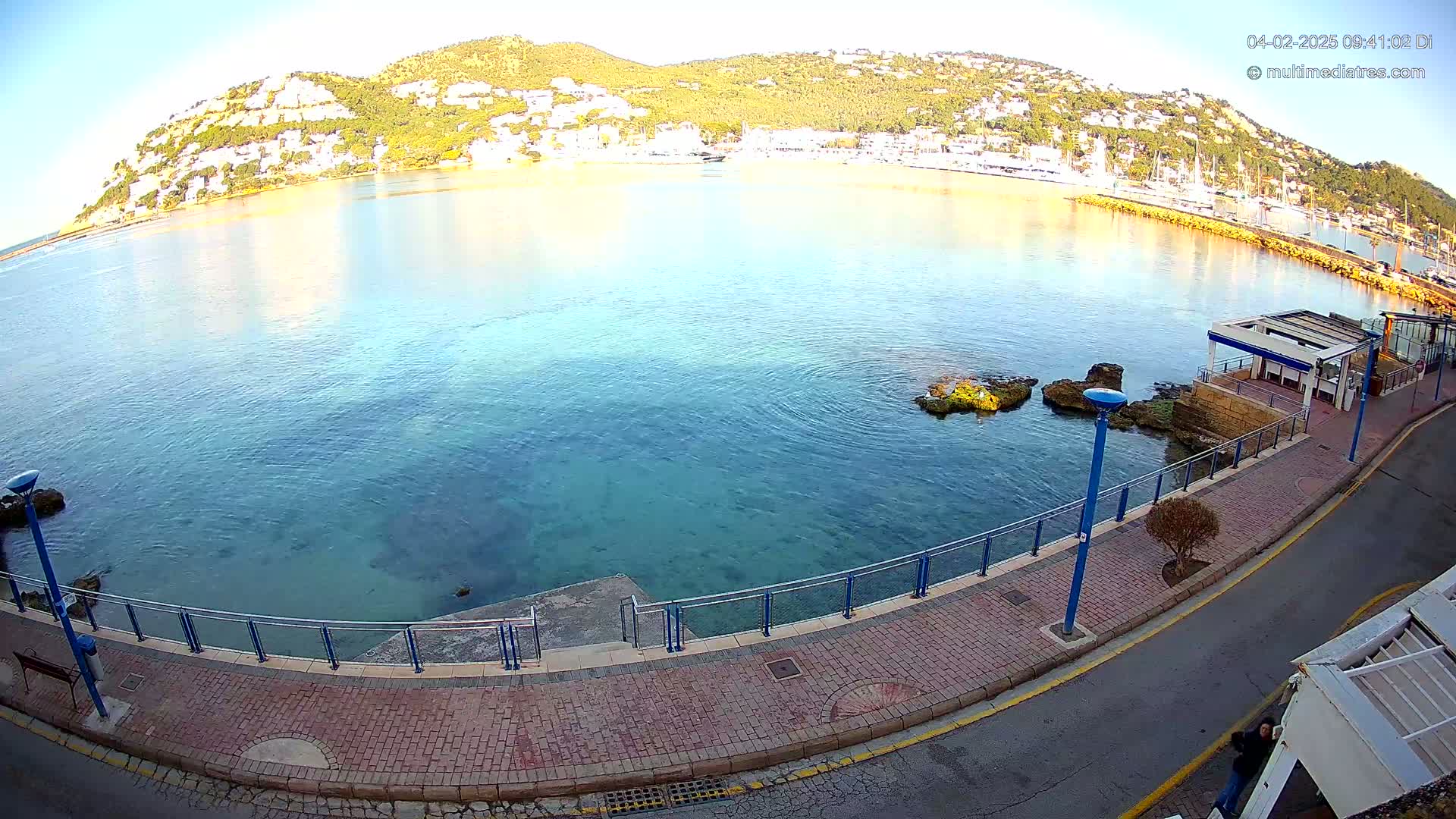 A calm, clear body of water laps a paved waterfront walkway next to a hill dotted with buildings under a bright, sunny sky.