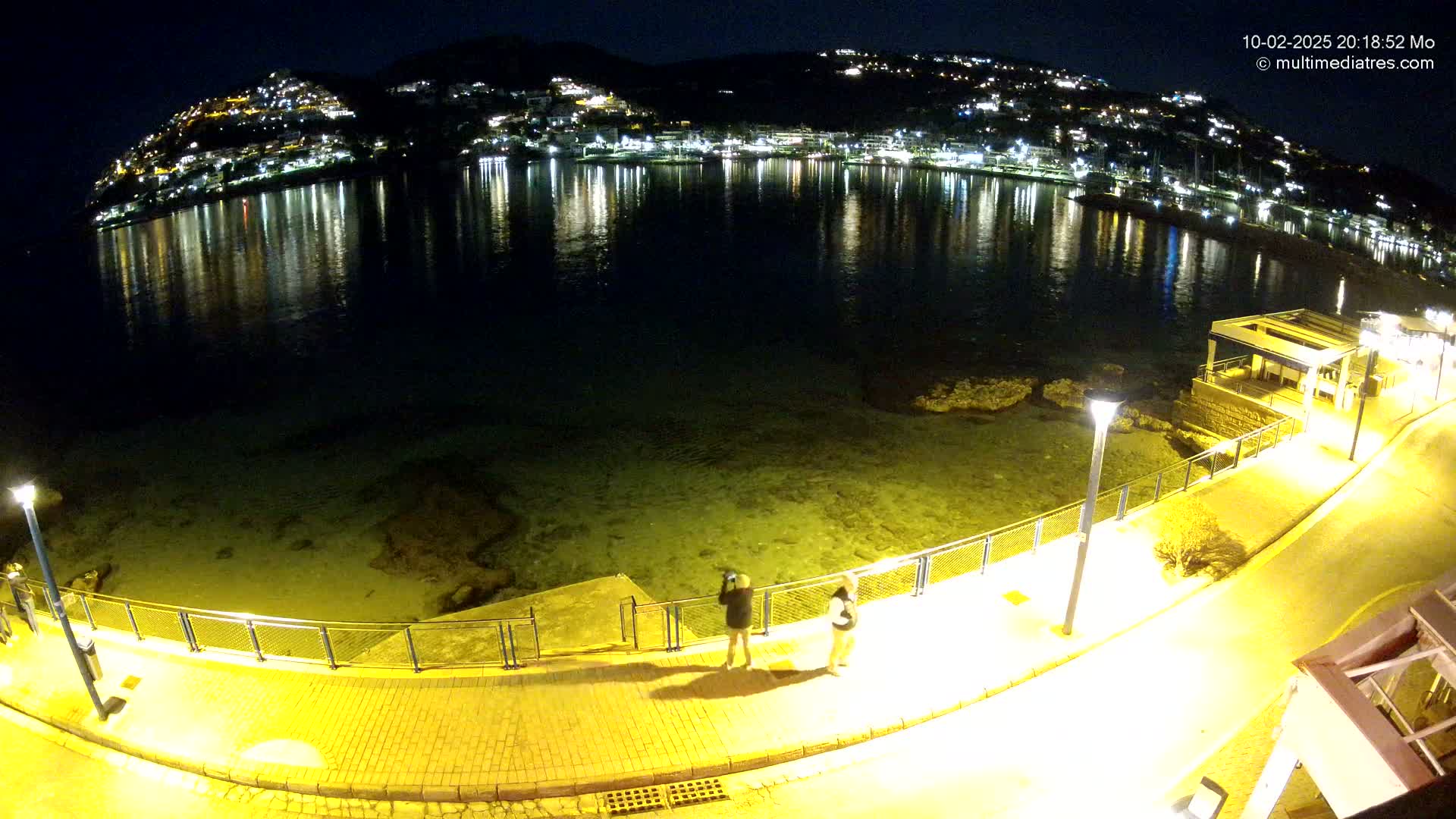 A nighttime, high-angle view shows a calm body of water reflecting the lights of a hillside town, with two people standing on a brightly lit waterfront path.