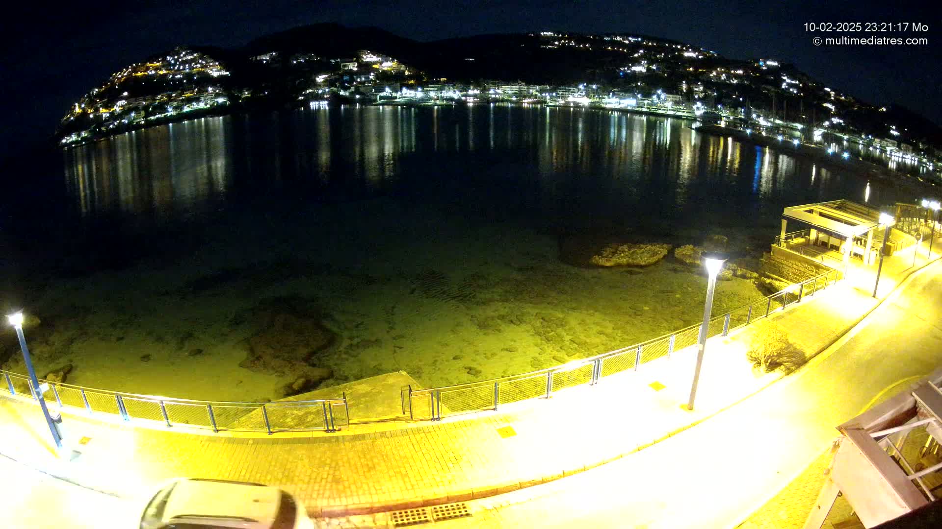 A nighttime coastal scene shows a calm body of water reflecting the lights of a town built on a hillside, with a paved walkway and lampposts along the shore.