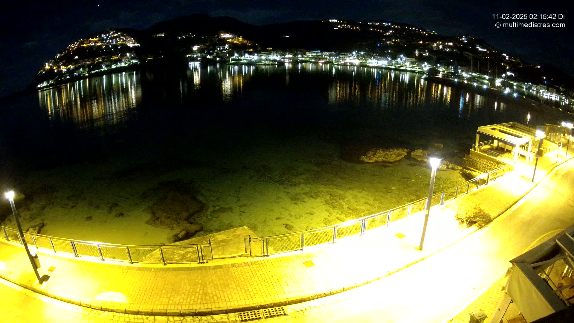 A nighttime view of a calm bay with a town's lights reflected in the water, illuminated walkways, and a small building near the shore.