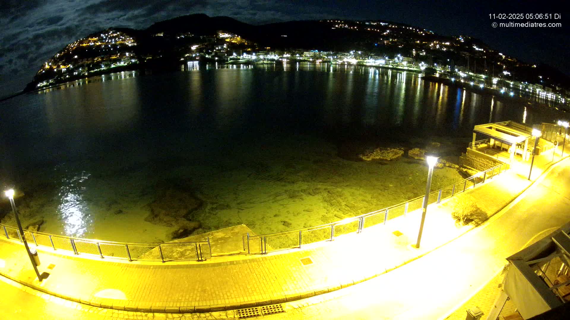 A nighttime, illuminated waterfront scene shows a calm body of water reflecting the lights of a hillside town under a cloudy sky.