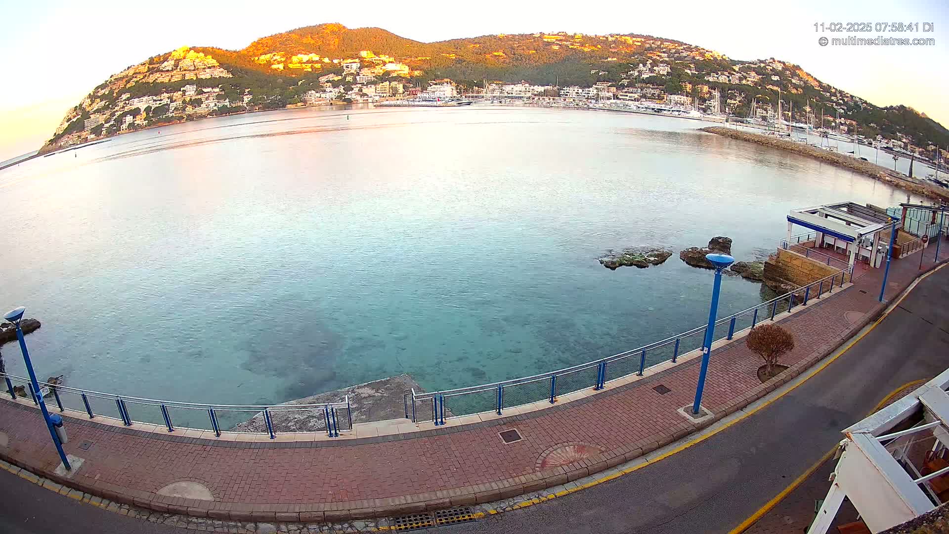 A calm, clear body of water laps a brick paved walkway beside a hill covered in buildings at sunrise.