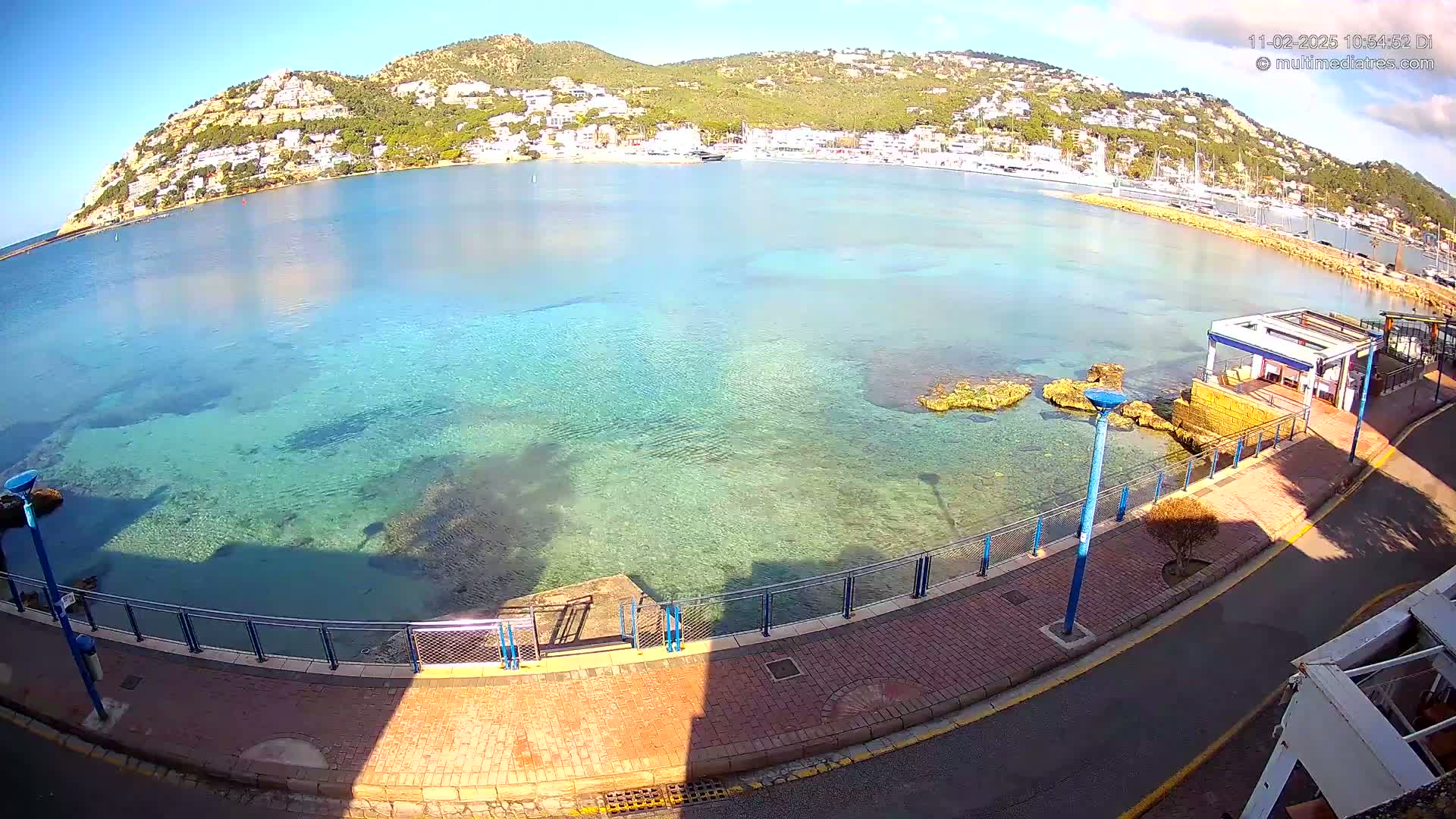 A clear, sunny day reveals a calm bay with crystal-clear turquoise water, a paved waterfront walkway, and a hillside town in the background.