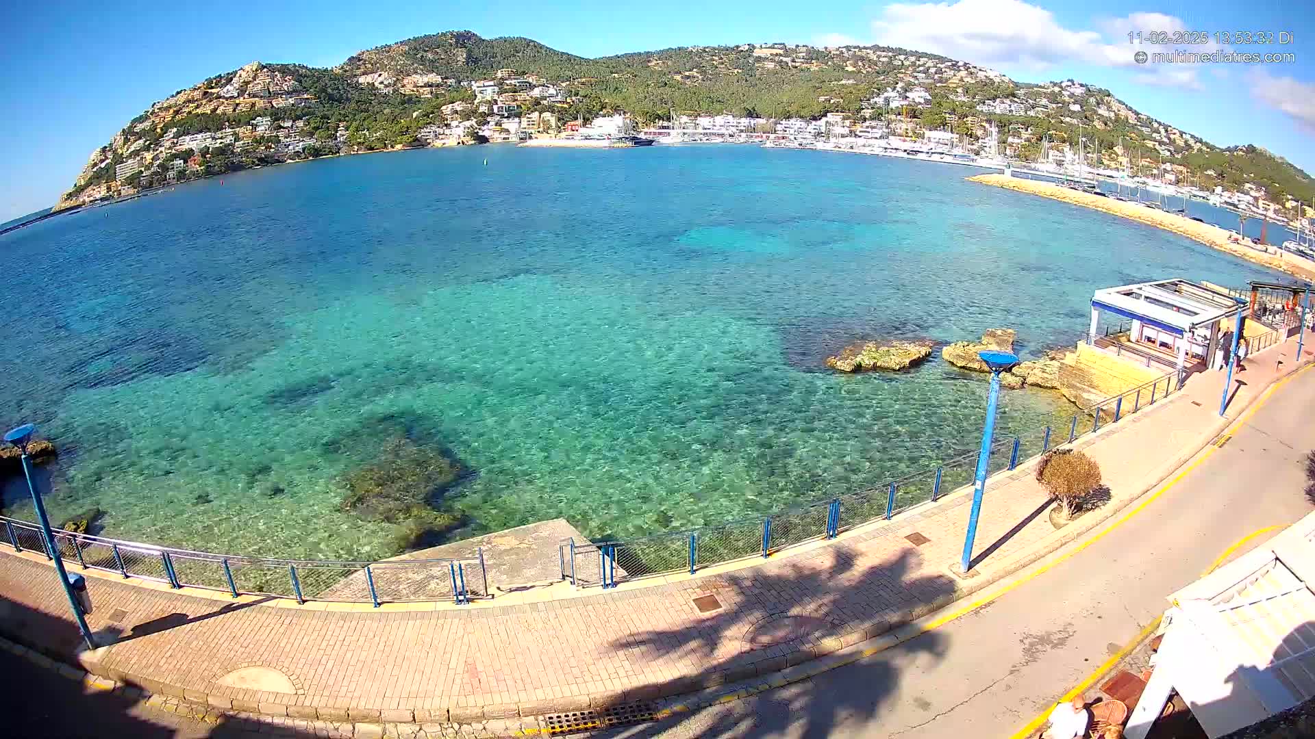 A clear, sunny day reveals a calm, shallow turquoise bay with a paved waterfront walkway and a small building; a hill covered with buildings and trees rises in the background.