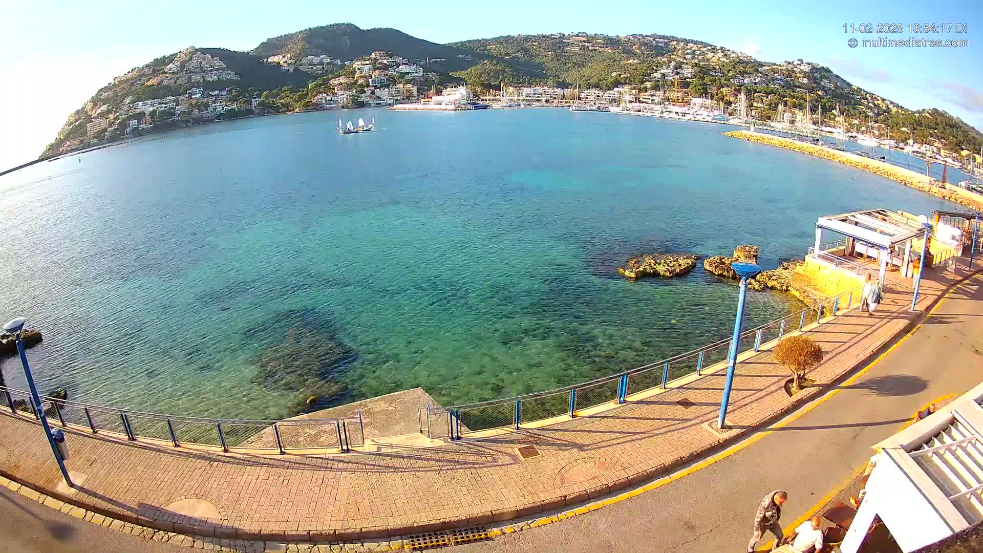 A clear, sunny day overlooks a calm, shallow, teal bay with a town and hills in the background, a paved walkway runs along the water's edge with a few people walking.