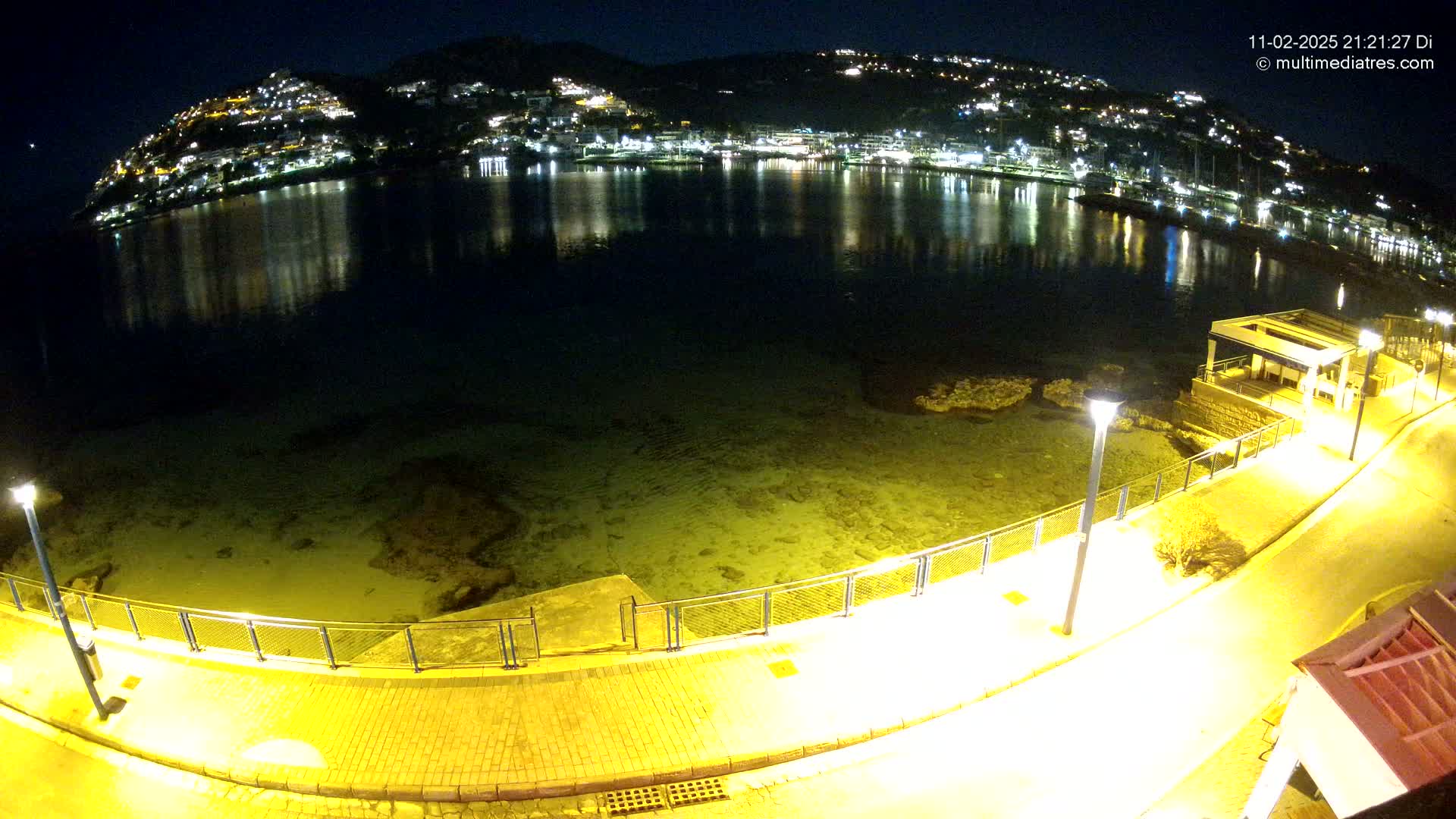 A nighttime view of a calm bay reflecting a hilltop town illuminated with lights, seen from a paved waterfront path with lamps.