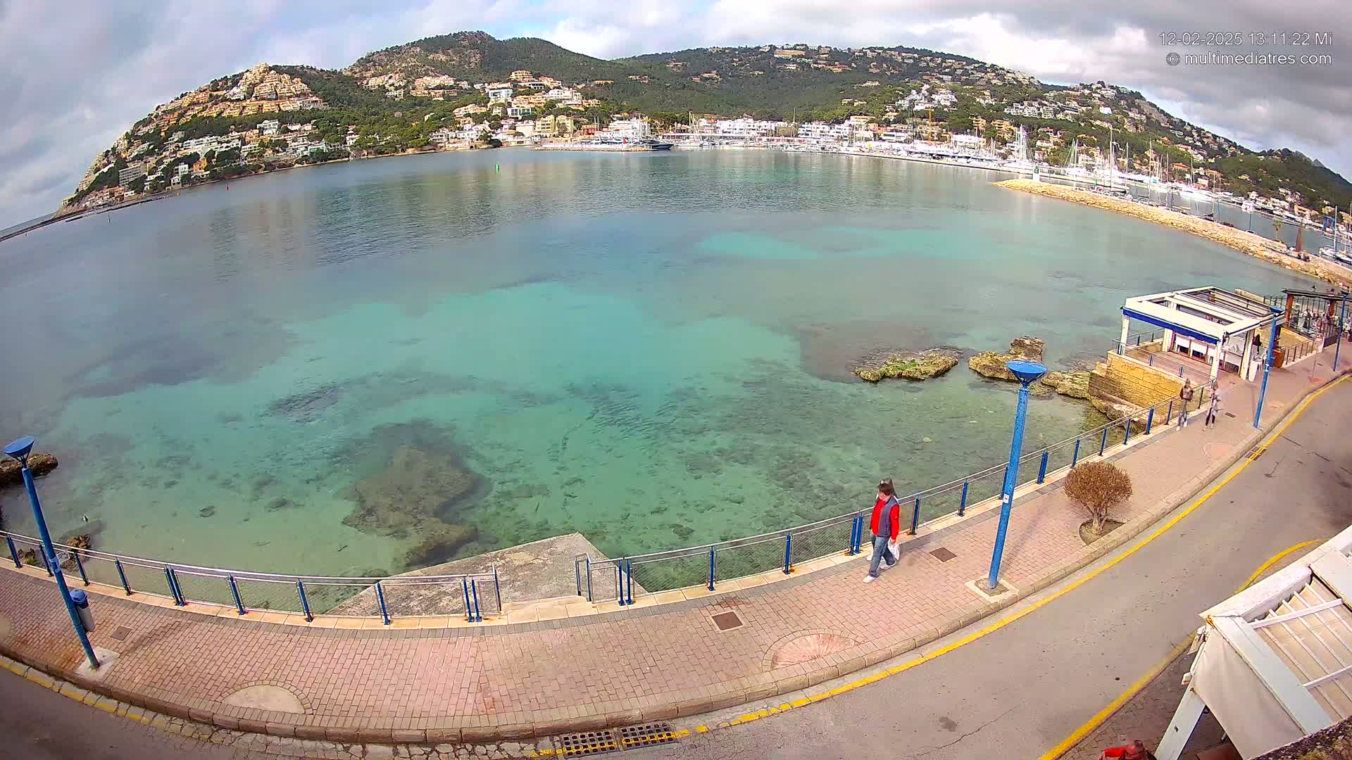 A person walks along a paved walkway overlooking a calm, clear turquoise bay with a town and hills in the background under a partly cloudy sky.