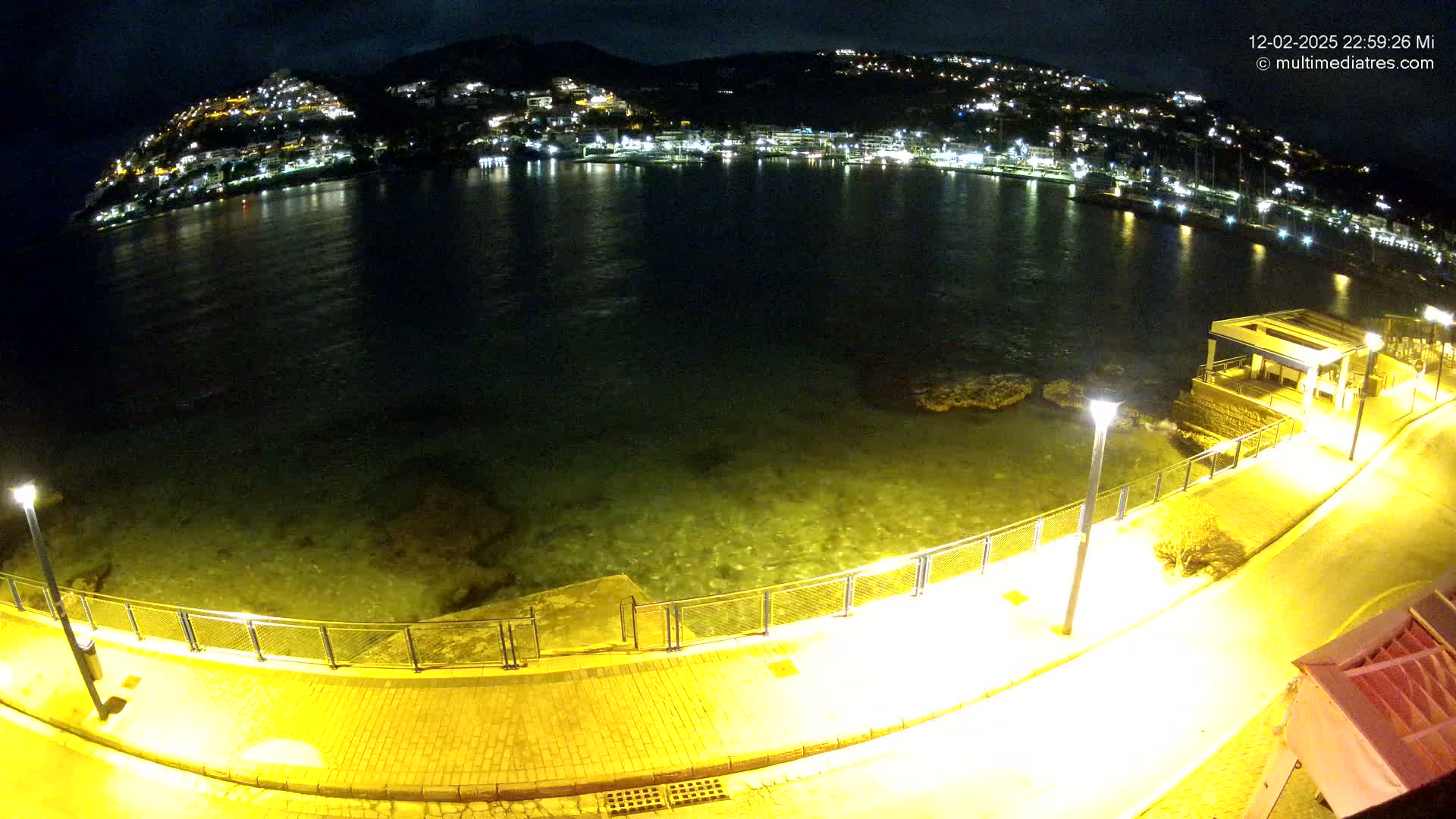A nighttime view of a calm, dark-colored body of water with a brightly lit town on a hillside across the water, a paved walkway along the shore, and a few illuminated lampposts.
