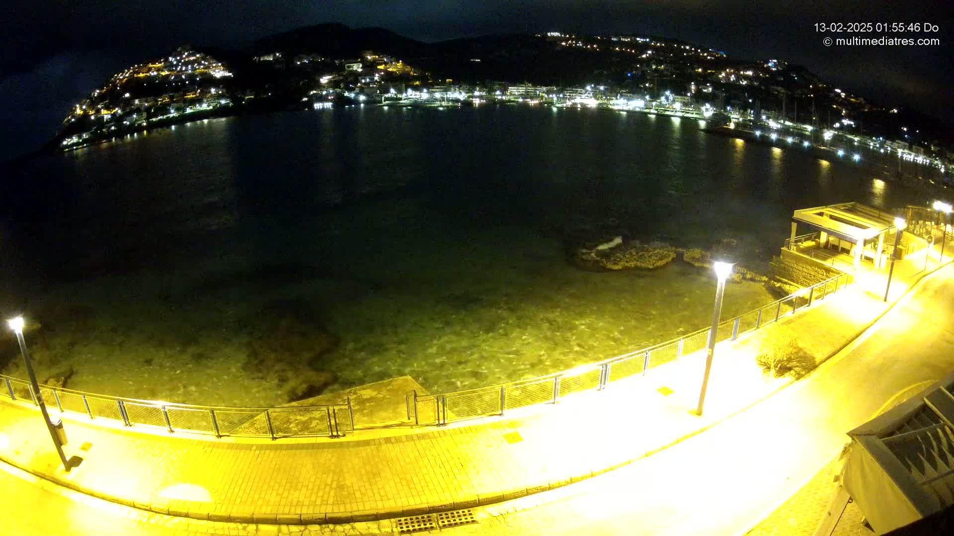 A nighttime view of a calm, dark-green body of water next to a paved walkway lined with lights, overlooking a hill dotted with illuminated buildings.