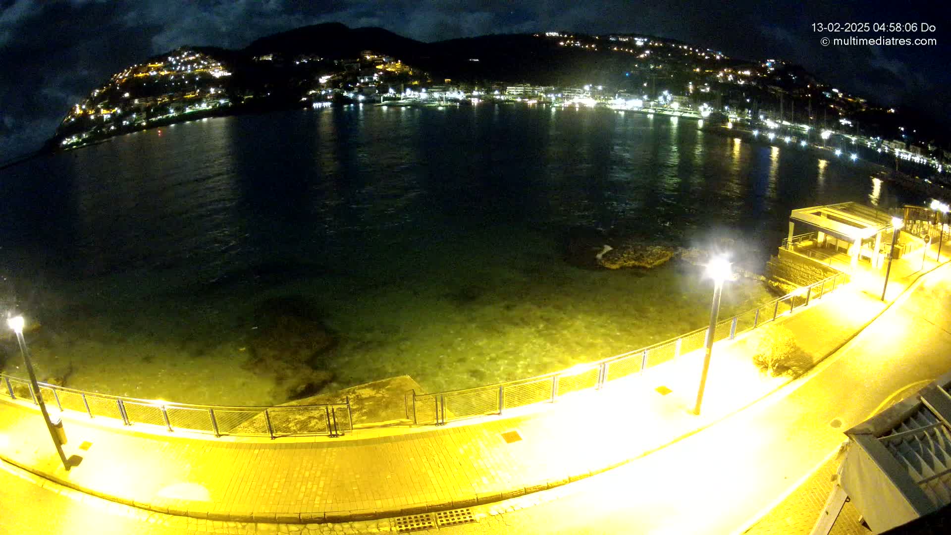 A nighttime, illuminated waterfront path curves alongside a calm, shallow body of water reflecting the lights of a hillside town.