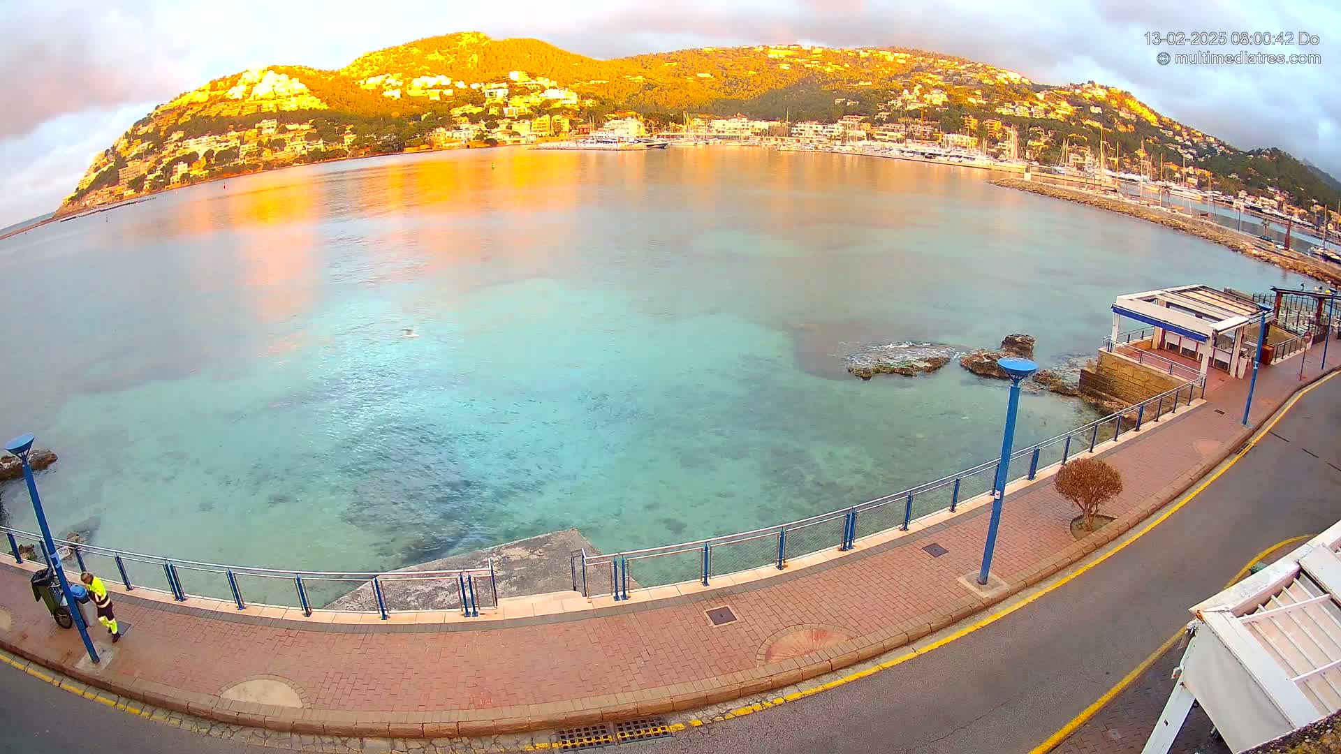 A calm, clear, teal-colored bay with a town and golden hills in the background is seen from an elevated viewpoint on a paved walkway next to a road.