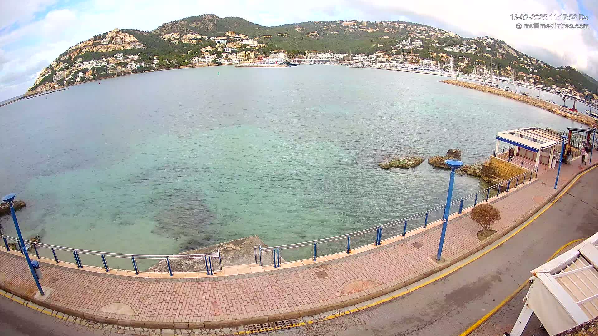 A paved walkway curves along a calm, clear, teal-colored bay, overlooking a marina filled with boats and a hillside town under a partly cloudy sky.