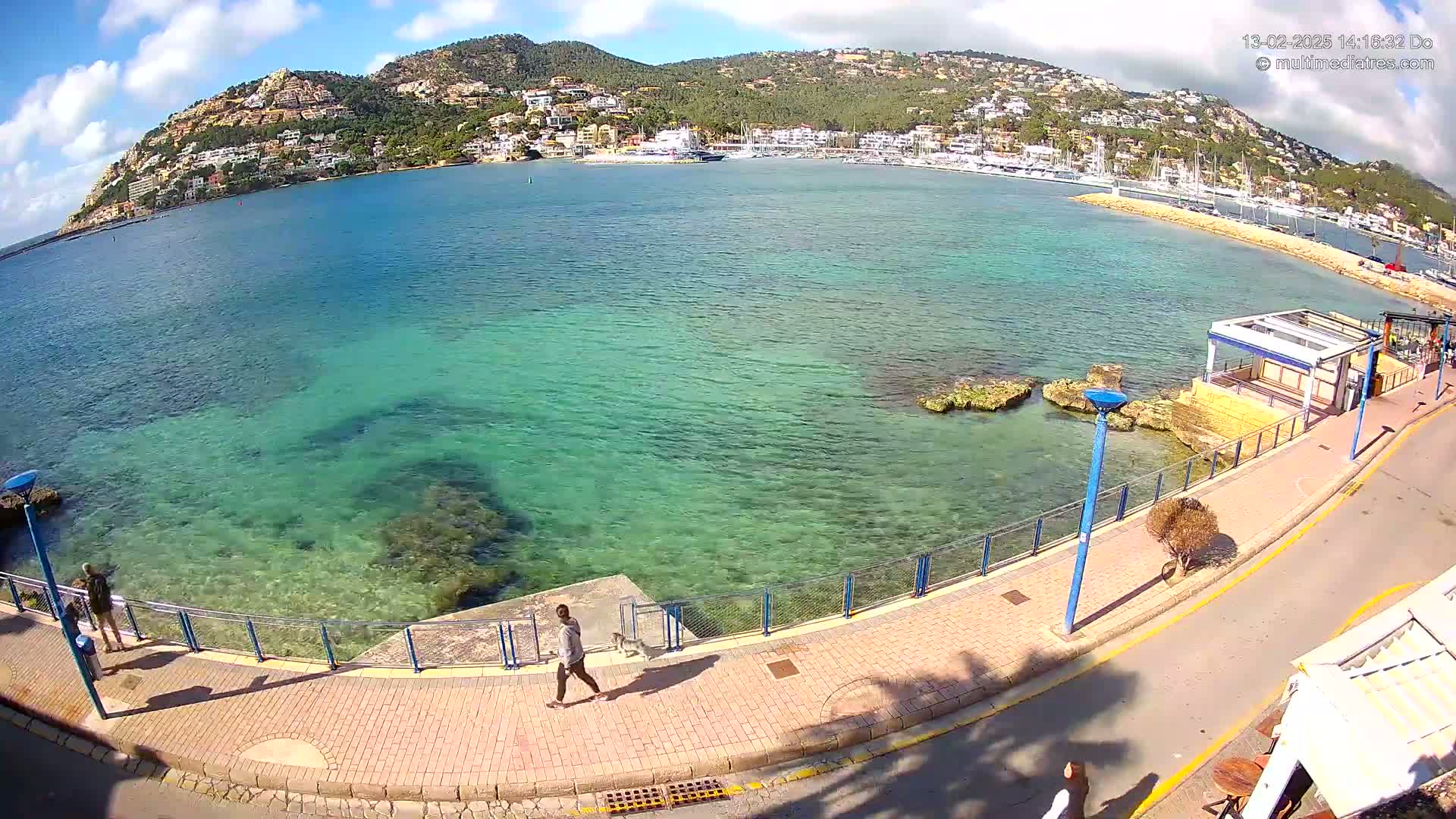 A person walks along a paved walkway next to a clear, teal-colored sea, with a hill covered in buildings and vegetation in the background under a partly sunny sky.
