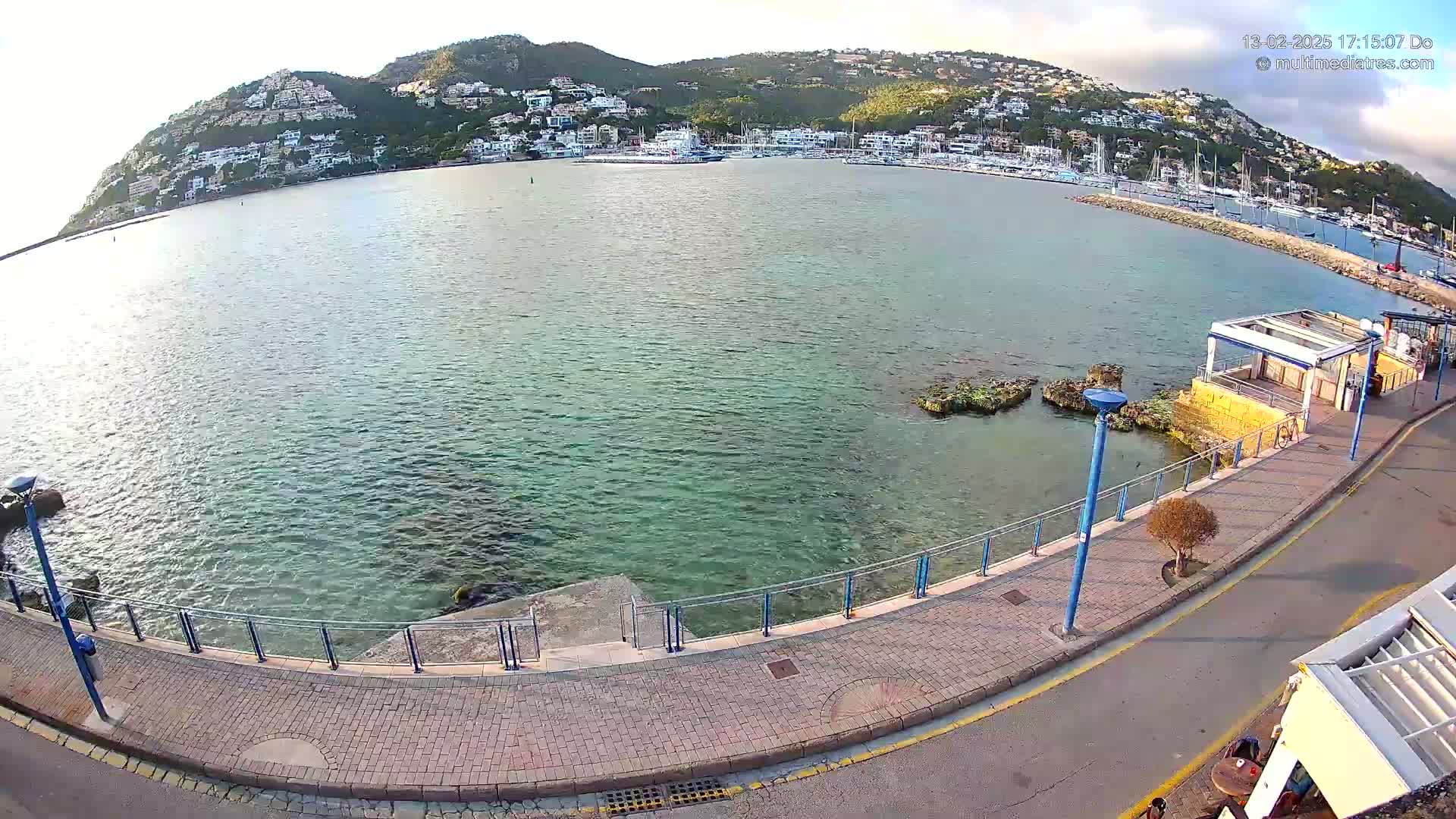 A calm, clear body of water laps against a paved waterfront walkway, with a hill covered in buildings visible in the background under a mostly clear sky.