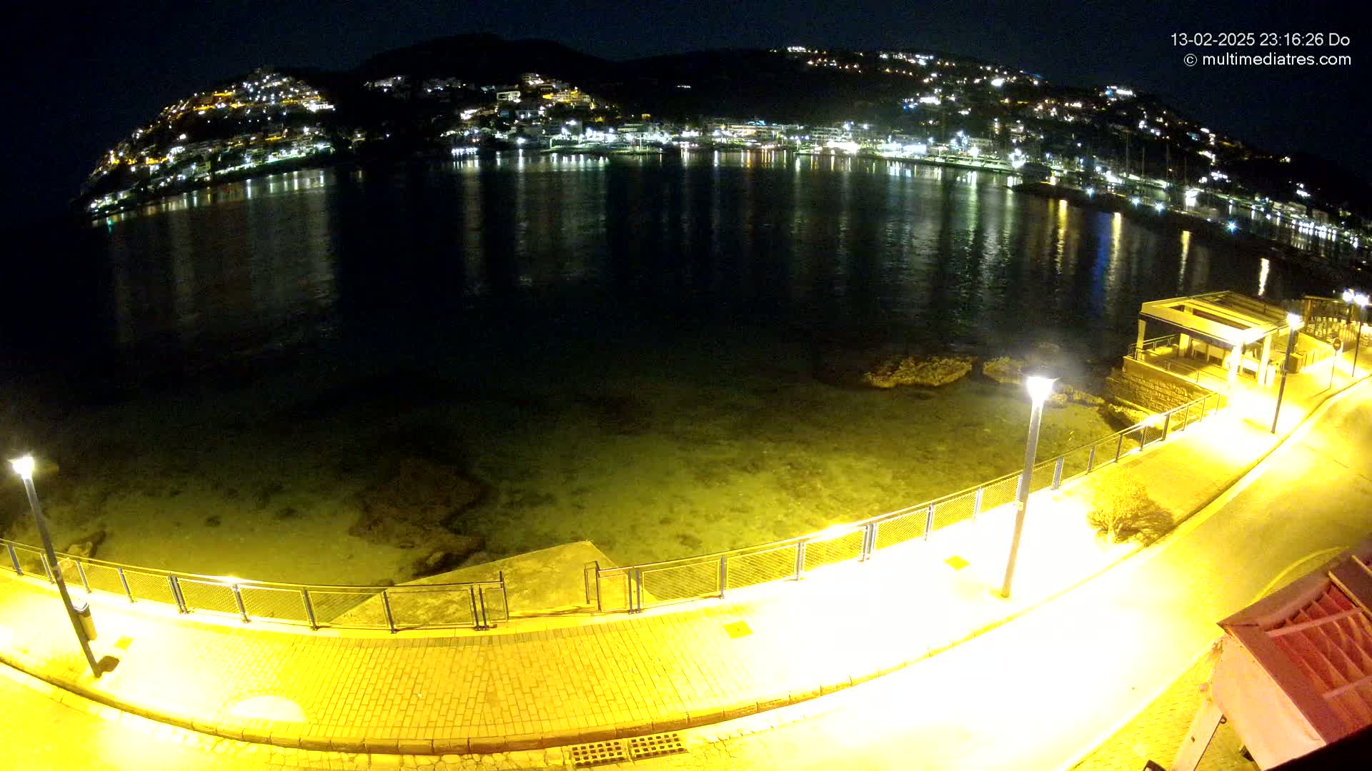 A nighttime view shows a calm body of water reflecting the lights of a town on a hillside, with a paved walkway and metal railing along the shoreline.