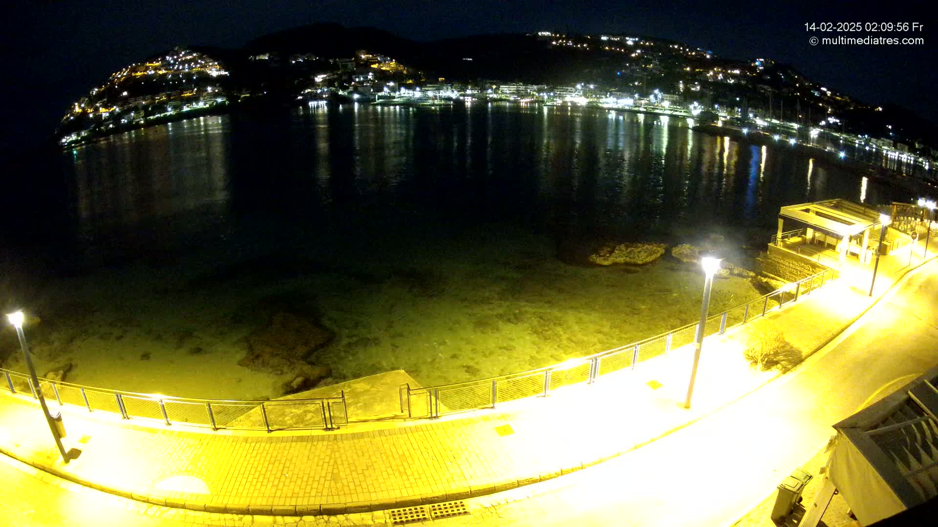 A nighttime view of a calm, shallow bay reflecting the lights of a hillside town, with a paved walkway and lampposts along the waterfront.
