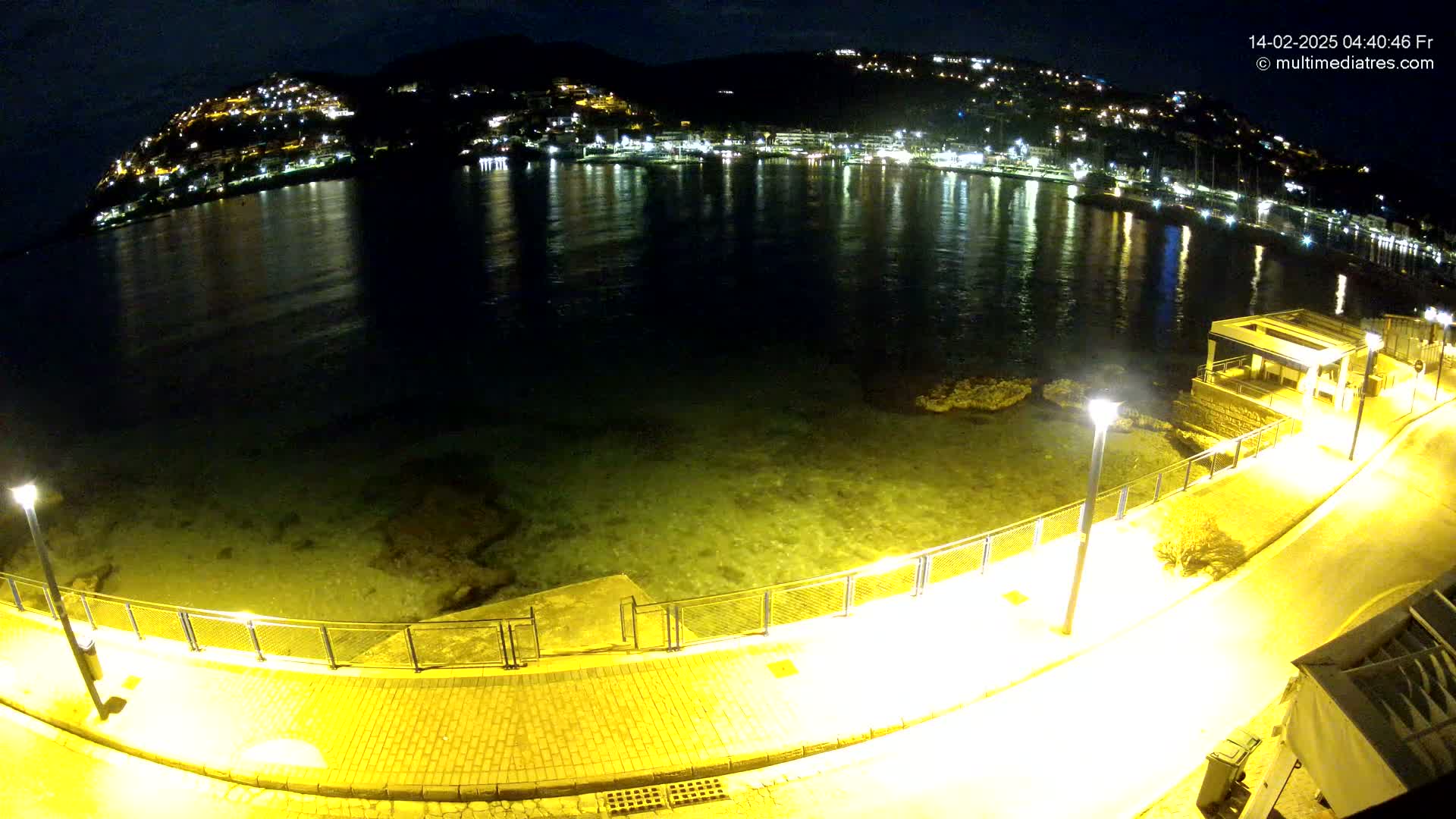 A nighttime view of a calm body of water reflecting the lights of a town nestled on a hillside, with a paved walkway and lampposts along the shore.