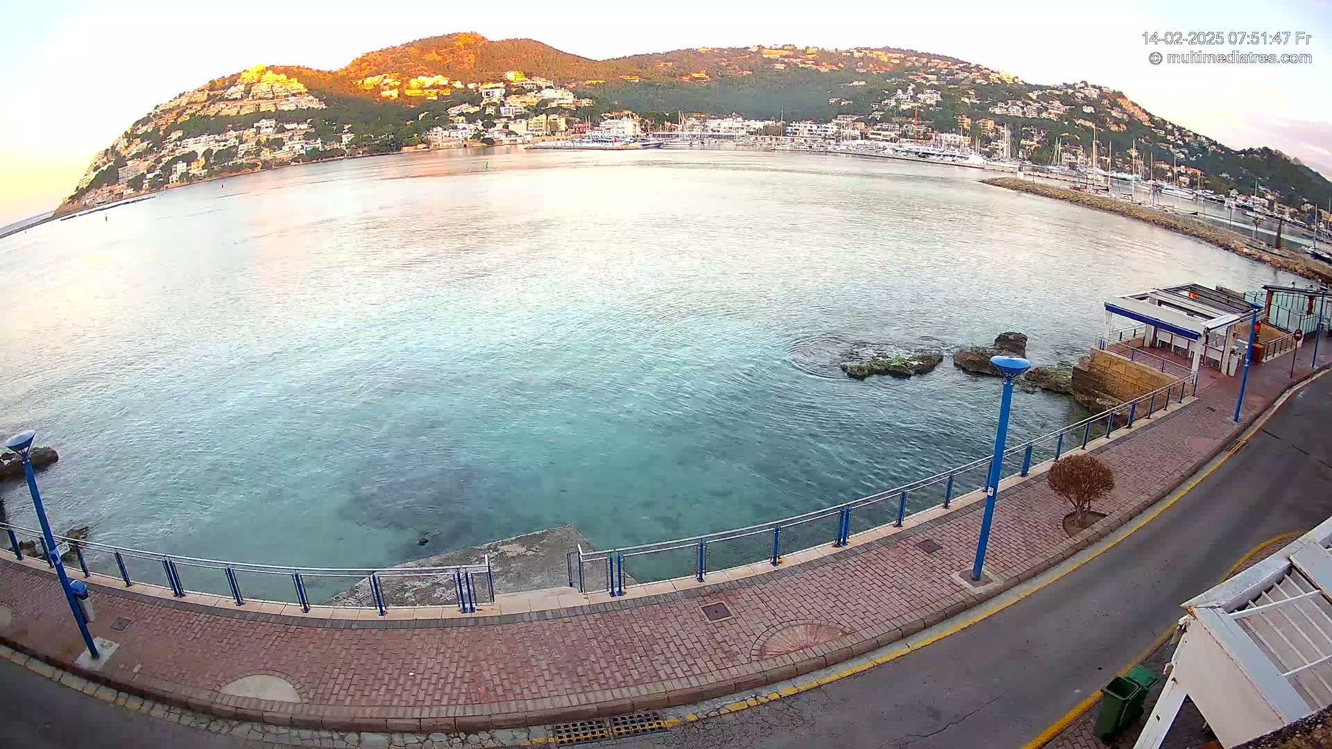 A calm, clear body of water laps a paved walkway beside a hillside town at sunrise.