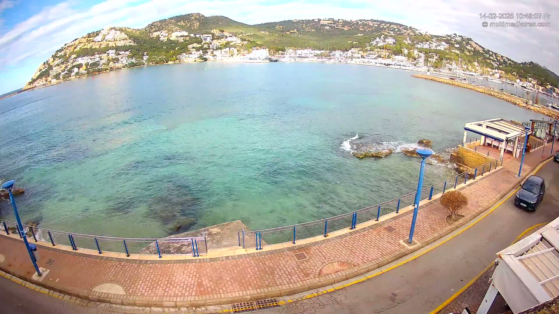A paved walkway lines a calm, clear turquoise bay, with a hill covered in buildings and trees in the background under a partly cloudy sky.