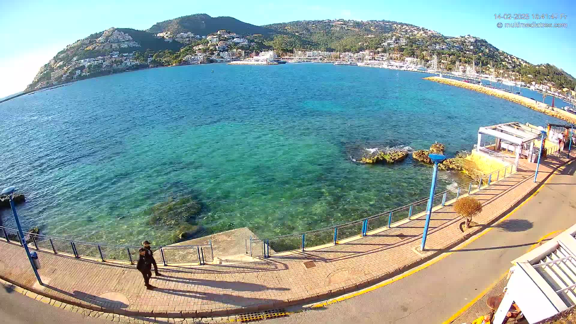 A clear, sunny day overlooks a calm, turquoise bay with a town and marina nestled against a hillside, as two people walk along a paved waterfront path.