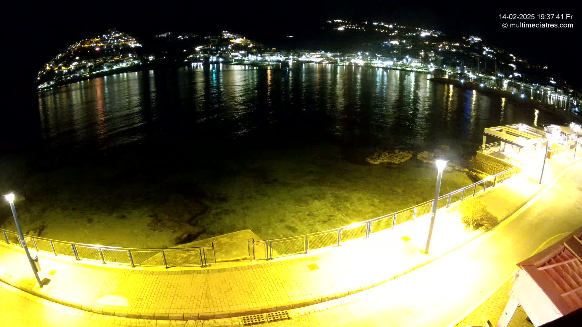 A nighttime view of a calm, dark-green body of water reflecting the lights of a hilltop town, seen from a paved waterfront walkway illuminated by streetlights.