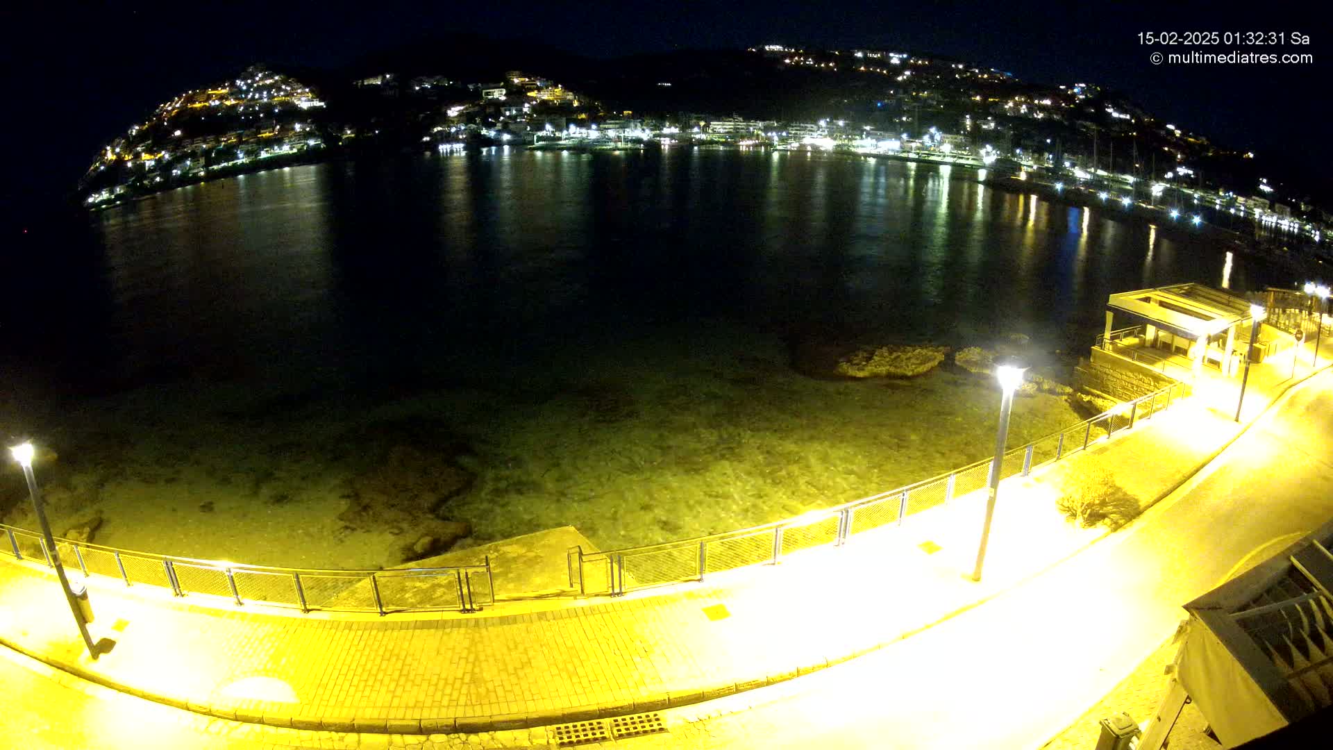 A nighttime view of a calm, shallow body of water with a brightly lit waterfront walkway and a hillside town in the background.
