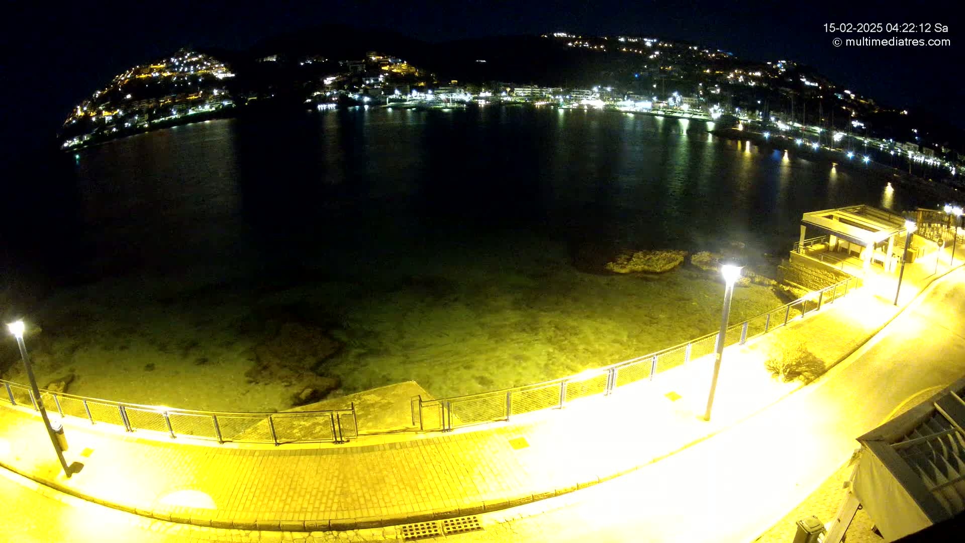 A nighttime view of a calm bay with a hilltop town illuminated across the water, a paved walkway alongside the water's edge, and several lampposts casting a yellow glow.